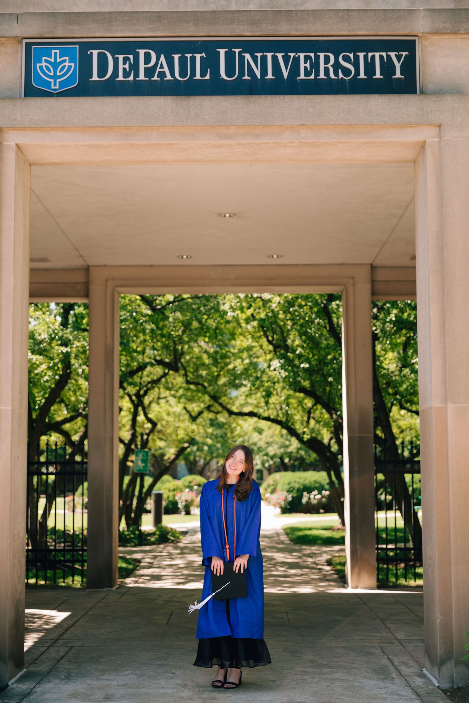 Graduate in blue cap and gown standing under DePaul University entrance archway surrounded by green trees.