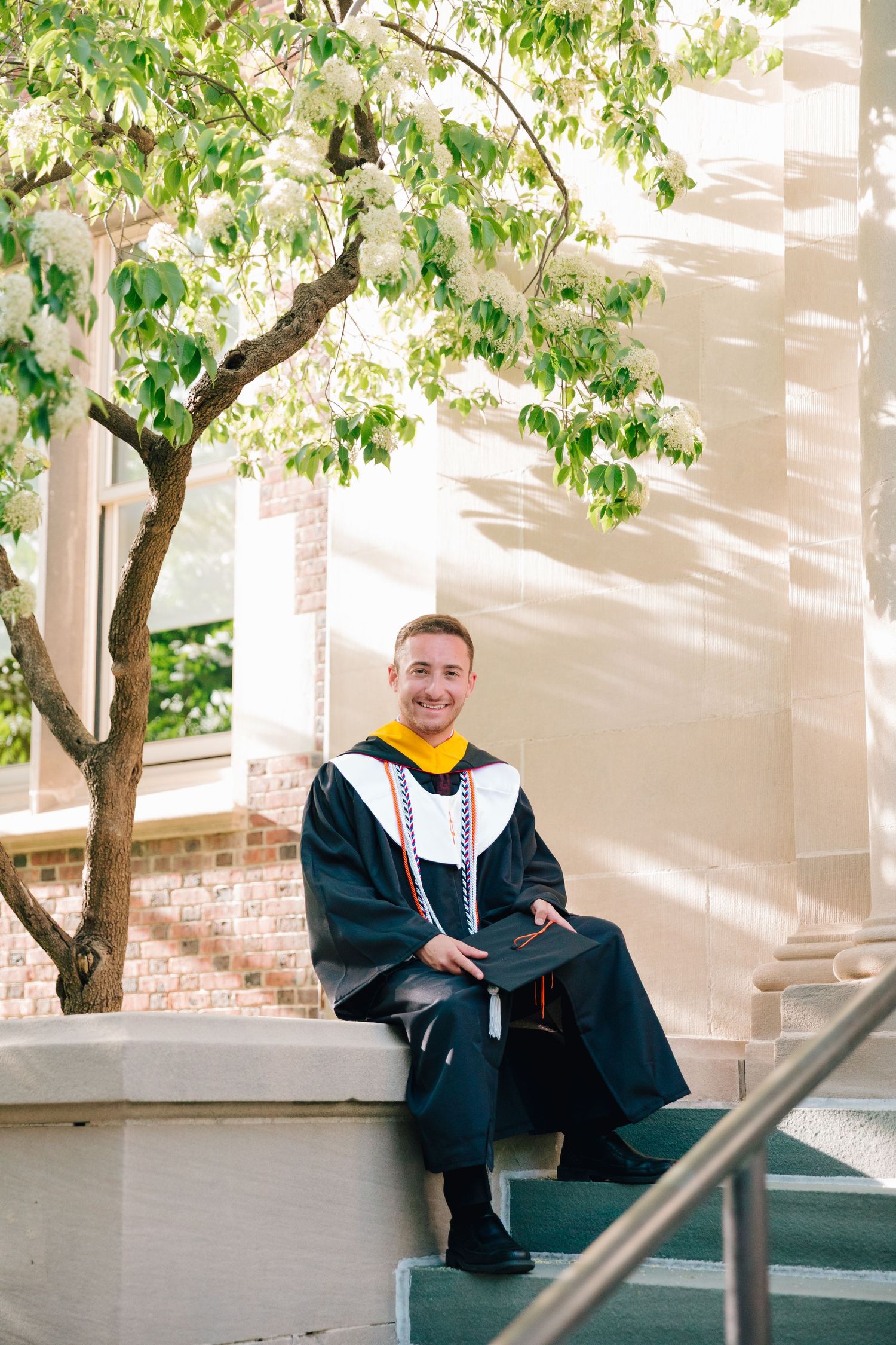 Graduate in dark academic regalia and yellow hood seated on outdoor stone steps.