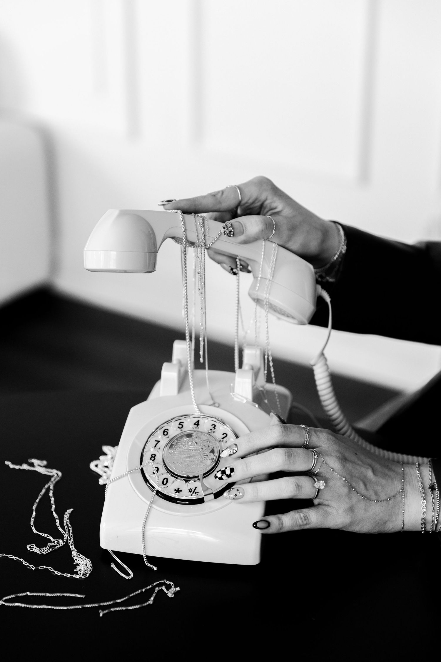 Black and white photo of hands with rings lifting a vintage rotary phone receiver with dangling necklaces on dark surface.