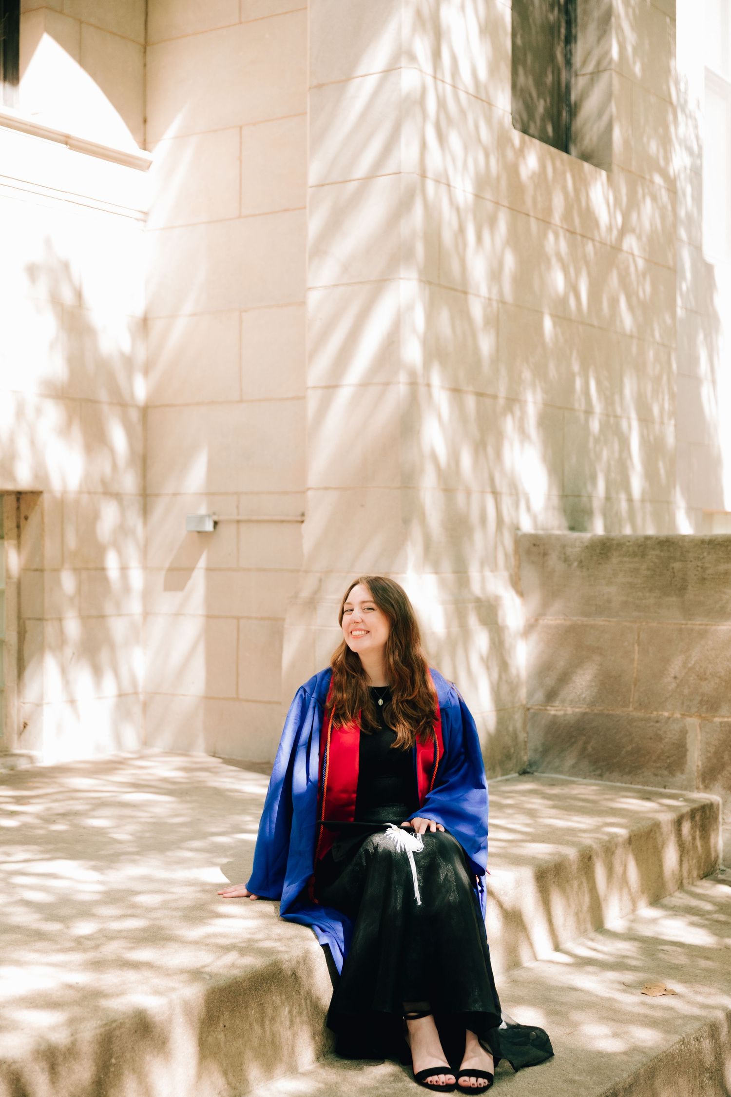 Graduate in blue commencement robe poses on sunlit stone steps with dappled shadows.