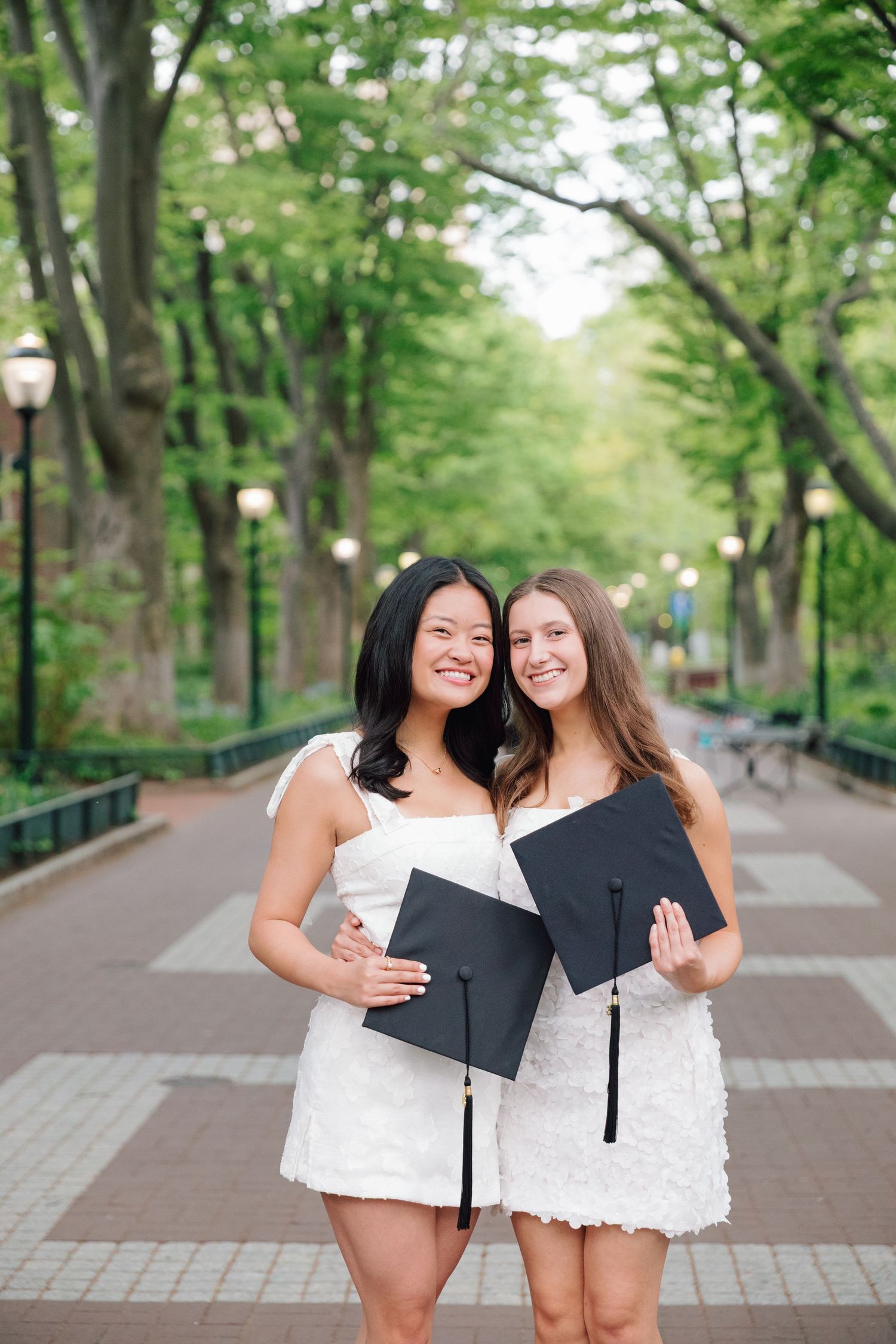 Two graduates in white dresses holding diplomas smile on tree-lined campus pathway.