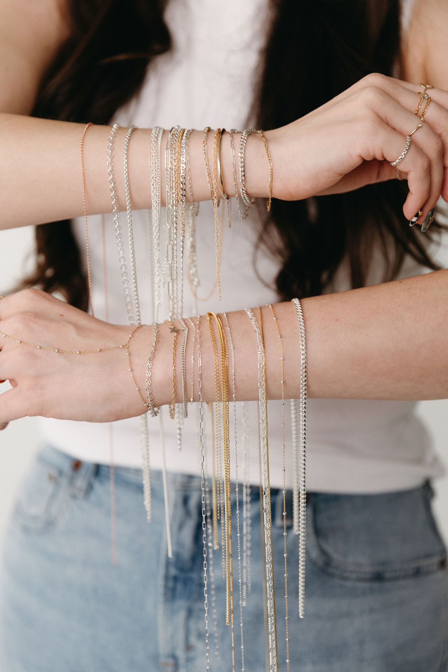 Woman displaying multiple gold and silver chain bracelets stacked on both wrists, wearing white top and jeans.
