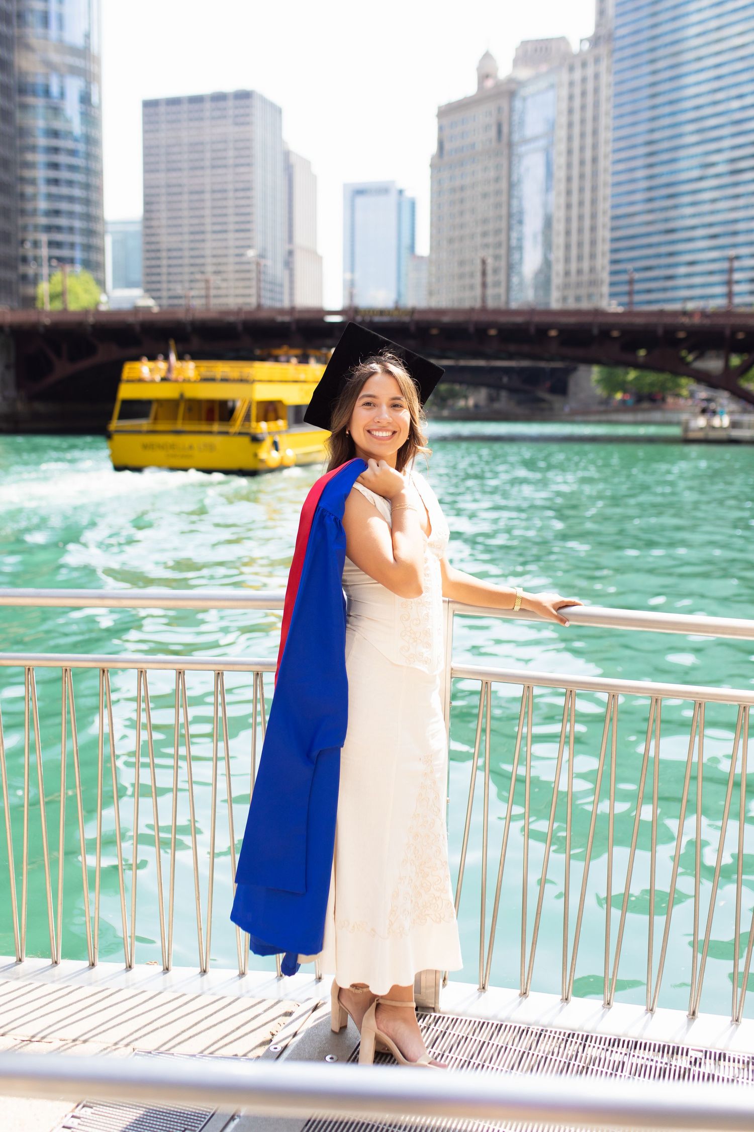 Graduate poses on Chicago riverwalk with city skyline and turquoise water in background.