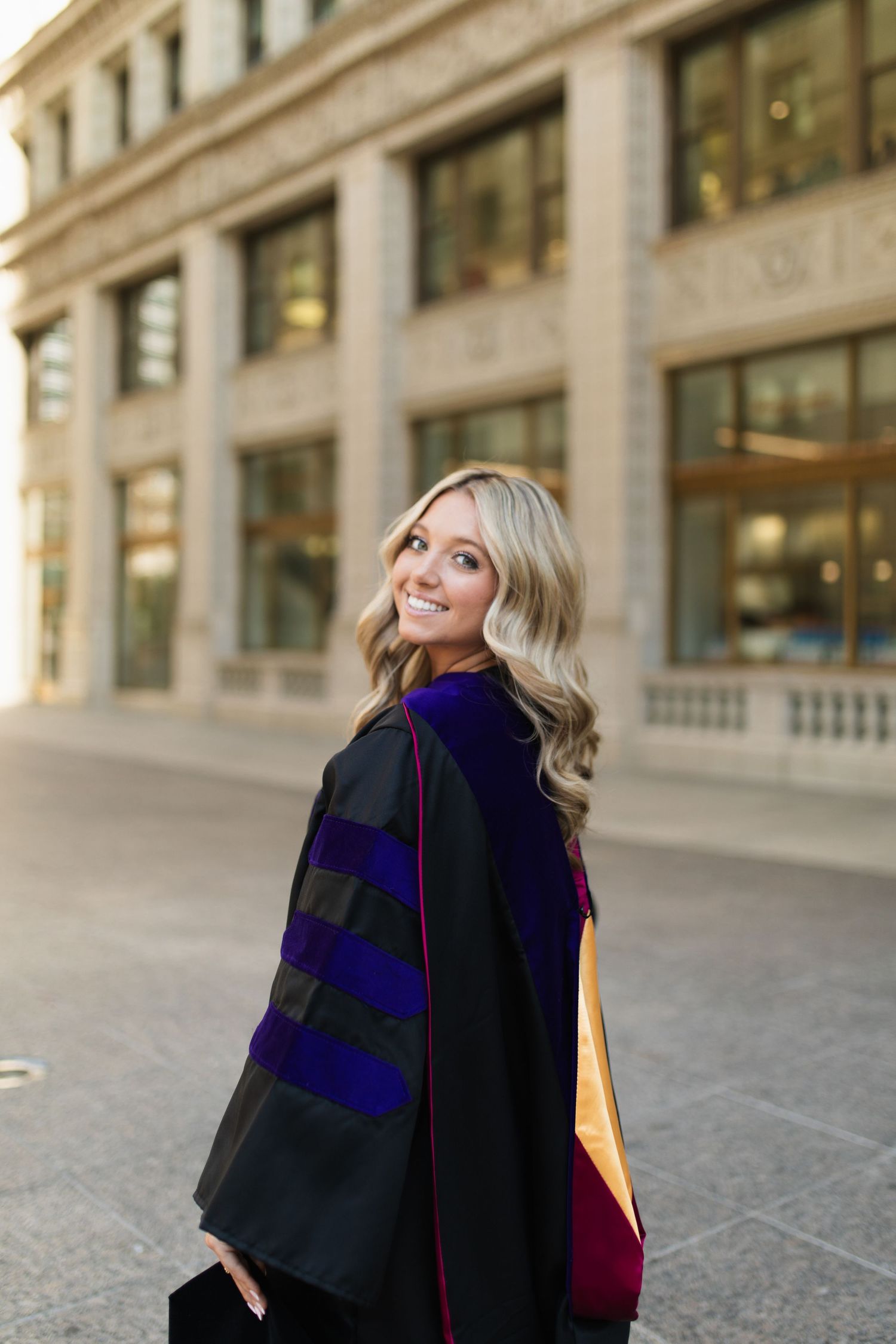 Graduate in academic regalia smiles over shoulder in front of historic building.