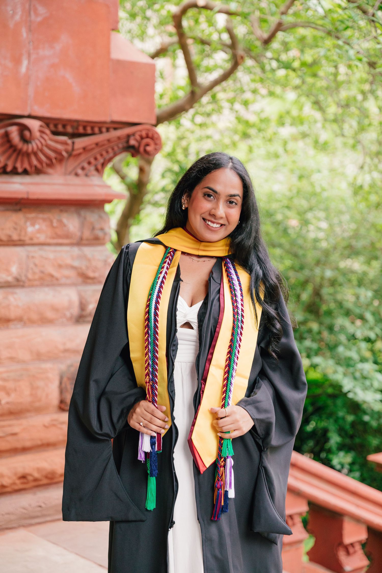 Graduate wearing colorful stole poses in academic regalia against brick wall.