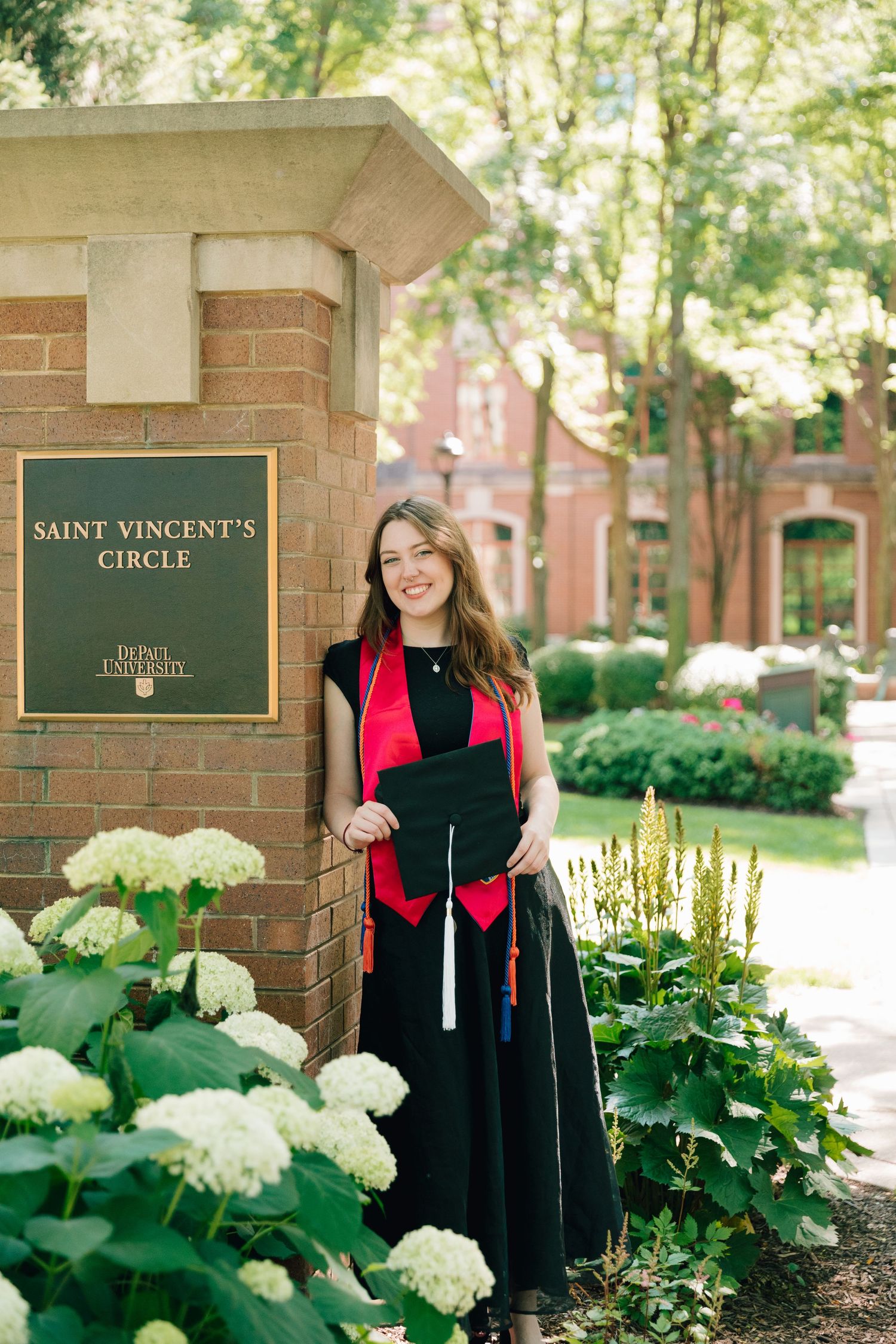 Graduate in black dress and red stole standing by Saint Vincent's Circle sign with blooming hydrangeas.