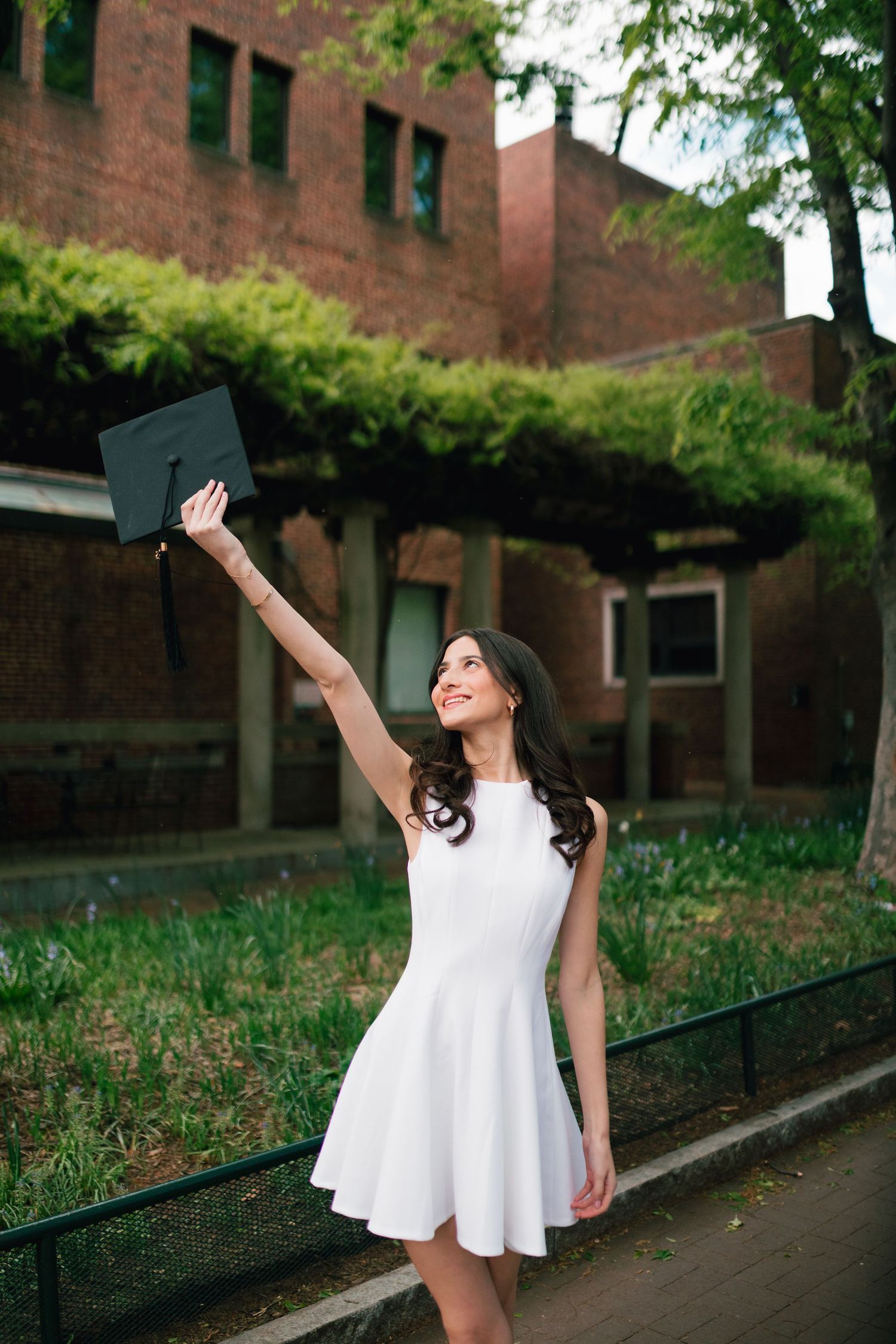 Graduate in white dress holds cap up joyfully on campus pathway.