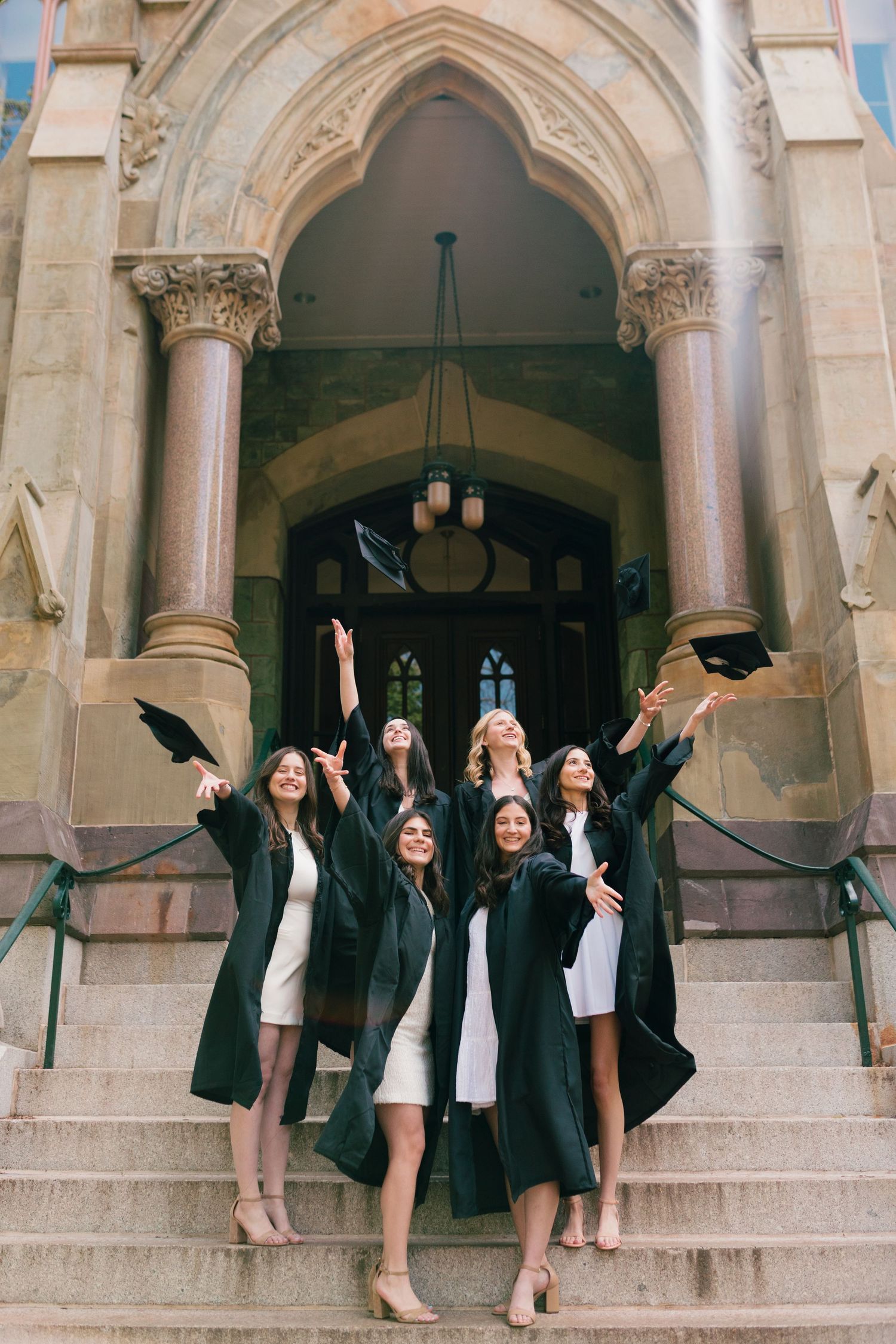 Graduates celebrate on stone steps in front of a Gothic-style archway entrance.