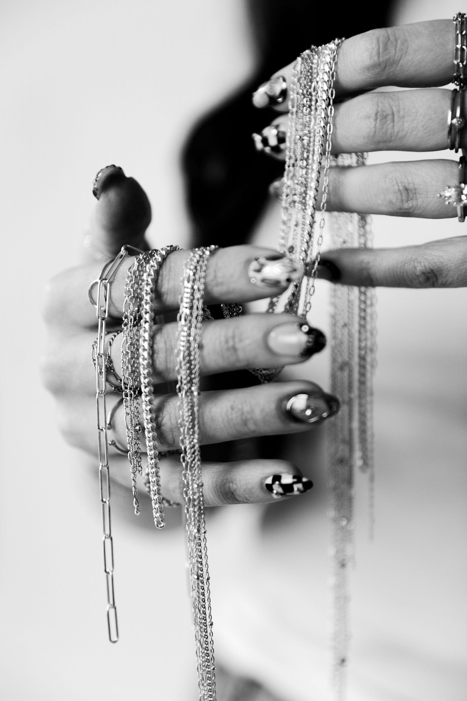 Black and white photo of hands adorned with multiple rings and necklace chains with decorative nail art.