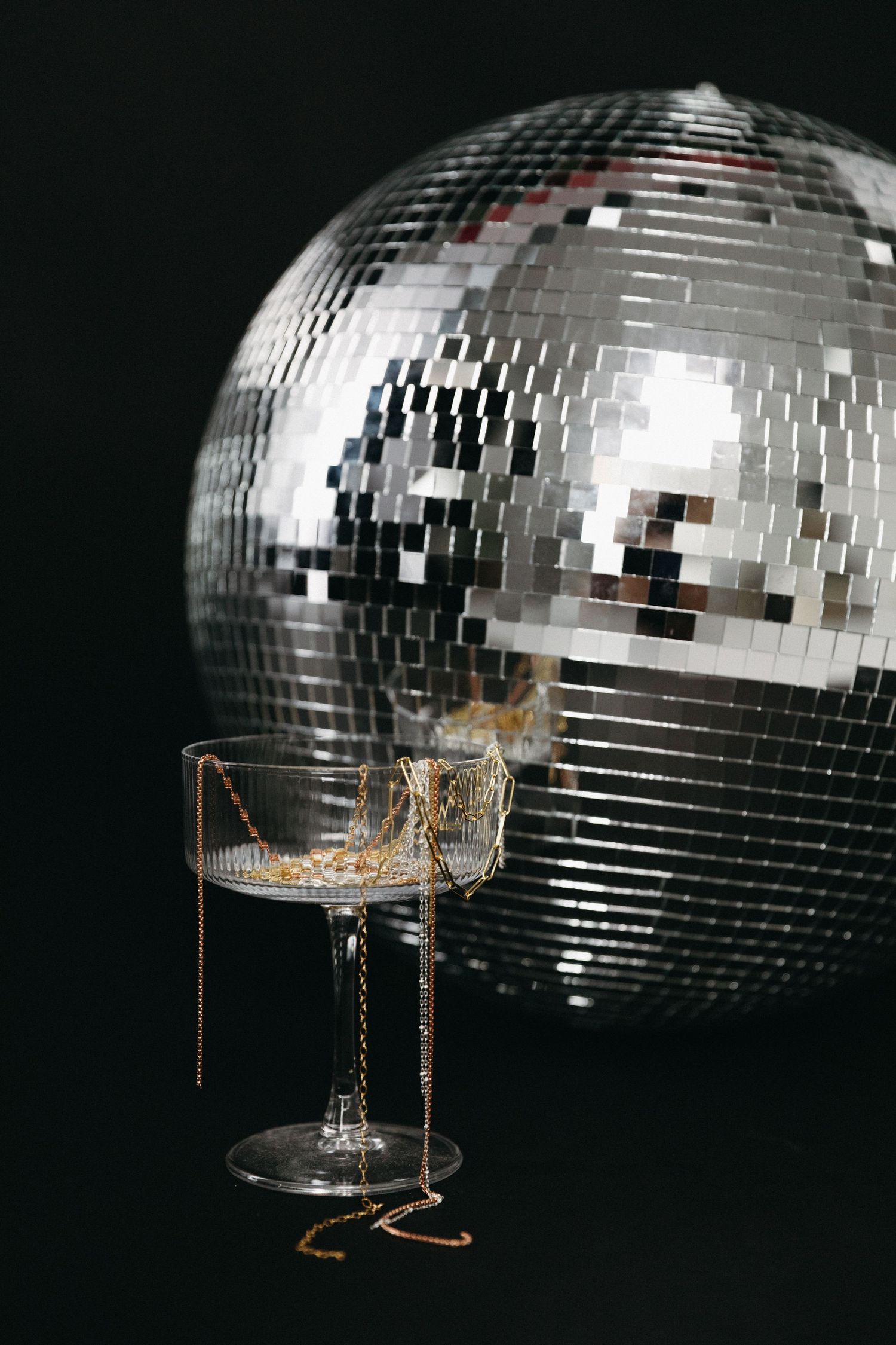 Sparkling disco ball with reflective mirror tiles behind a crystal champagne coupe glass with gold confetti on black background.