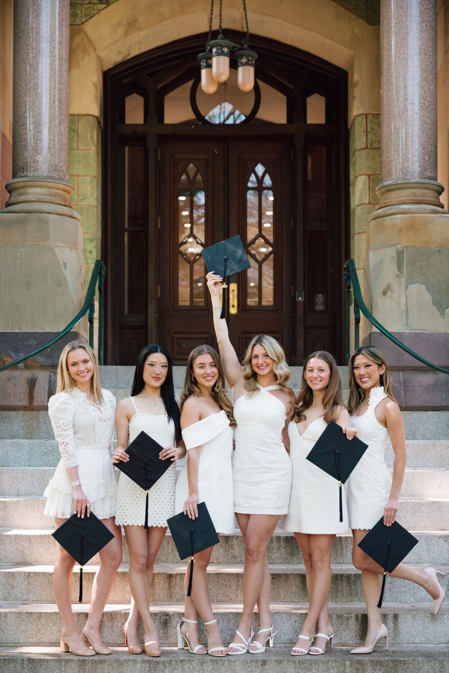 Graduates in white dresses holding diplomas pose on stone steps of gothic university building.