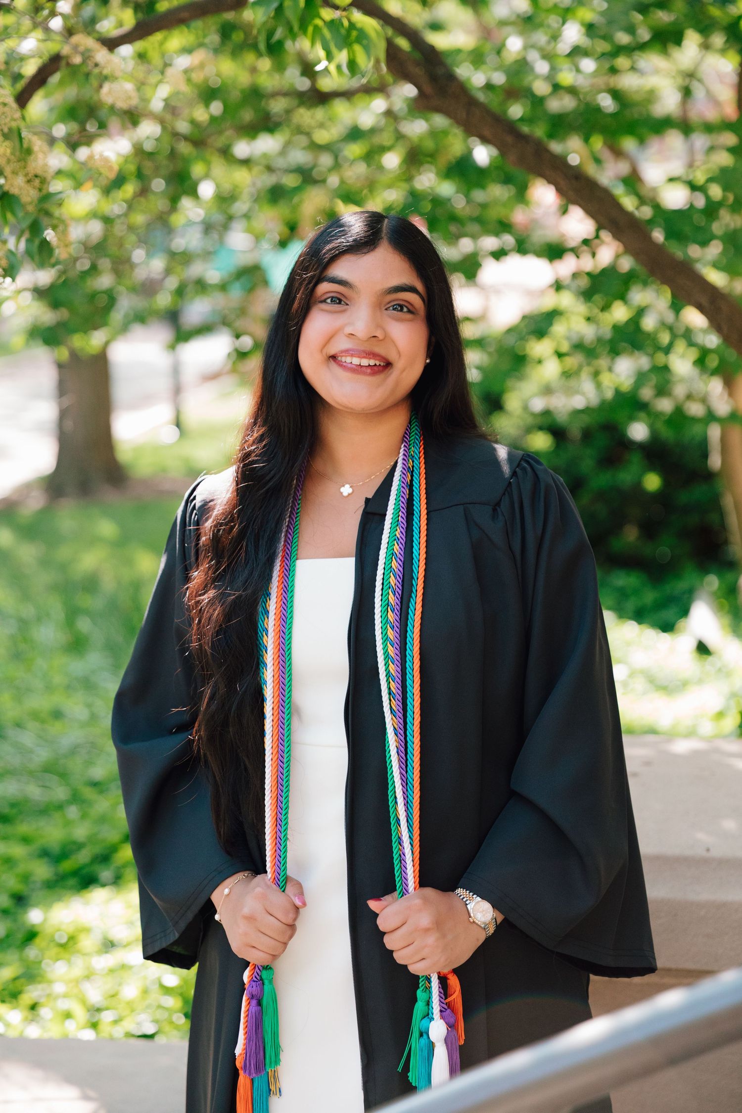 Graduate in black academic regalia with colorful stole smiling on tree-lined campus walkway.