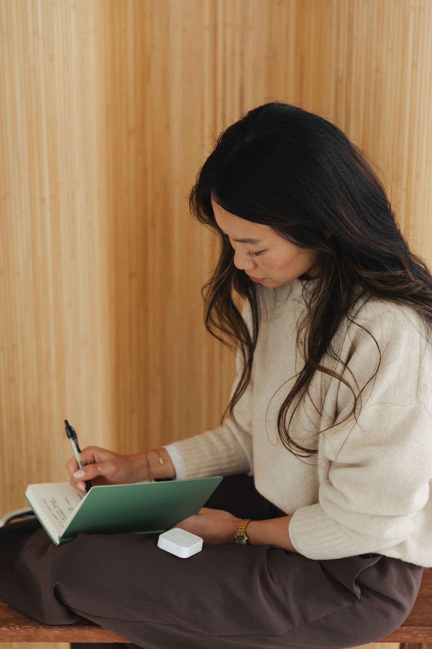Woman in cream sweater sitting cross-legged, writing in a green notebook against a wooden wall background.