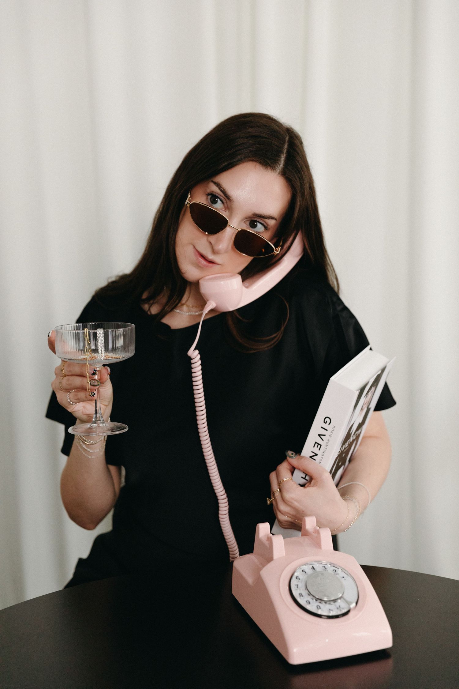 Woman in black dress and sunglasses holds wine glass while talking on pink retro rotary phone at dark table.