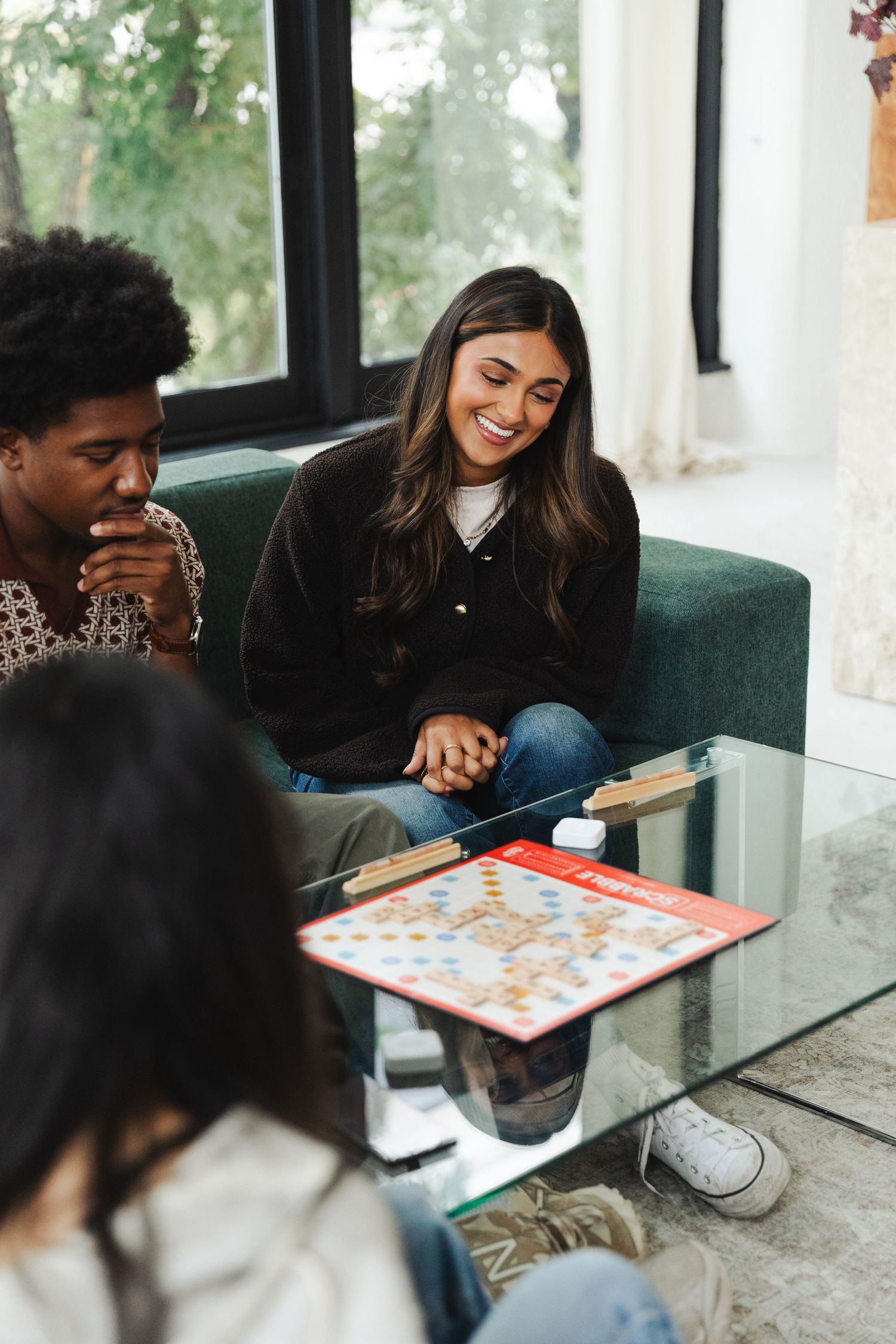 Friends laughing and playing Scrabble board game together on a glass coffee table in a bright living room.