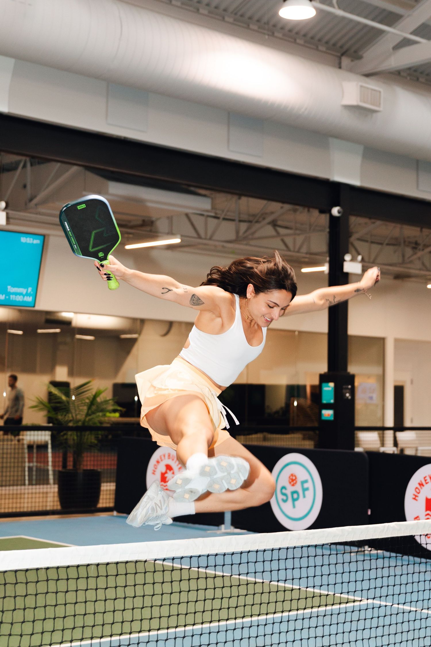 Woman leaping over pickleball net mid-game, holding green paddle in dynamic action shot at indoor SPF court.
