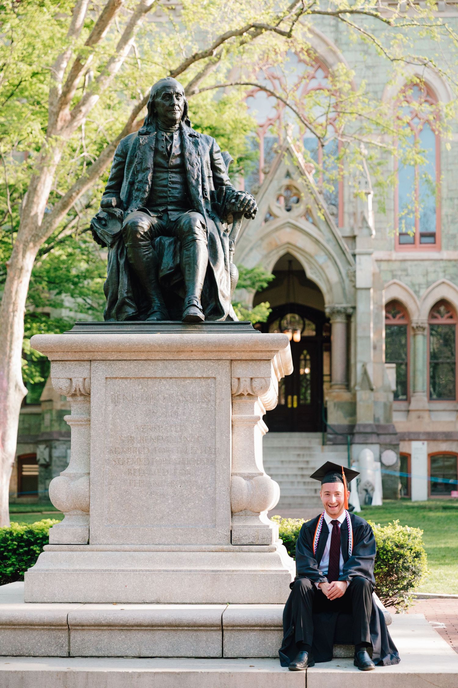 Graduate seated at base of Benjamin Franklin statue on university campus.