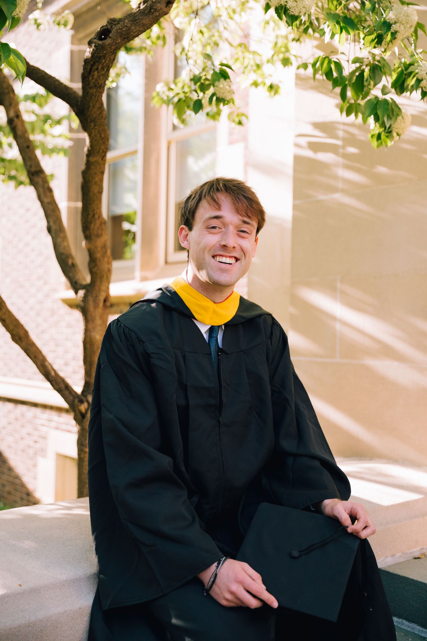 Person in graduation gown and yellow shirt poses cheerfully under tree branches.