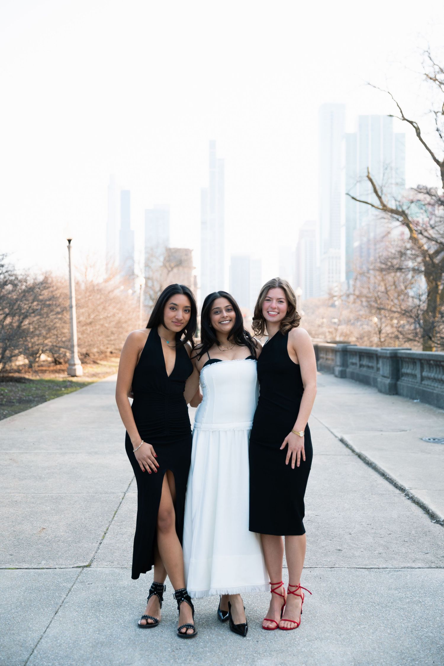 Three graduates in black and white dresses pose together on a sunny day with city buildings in background.