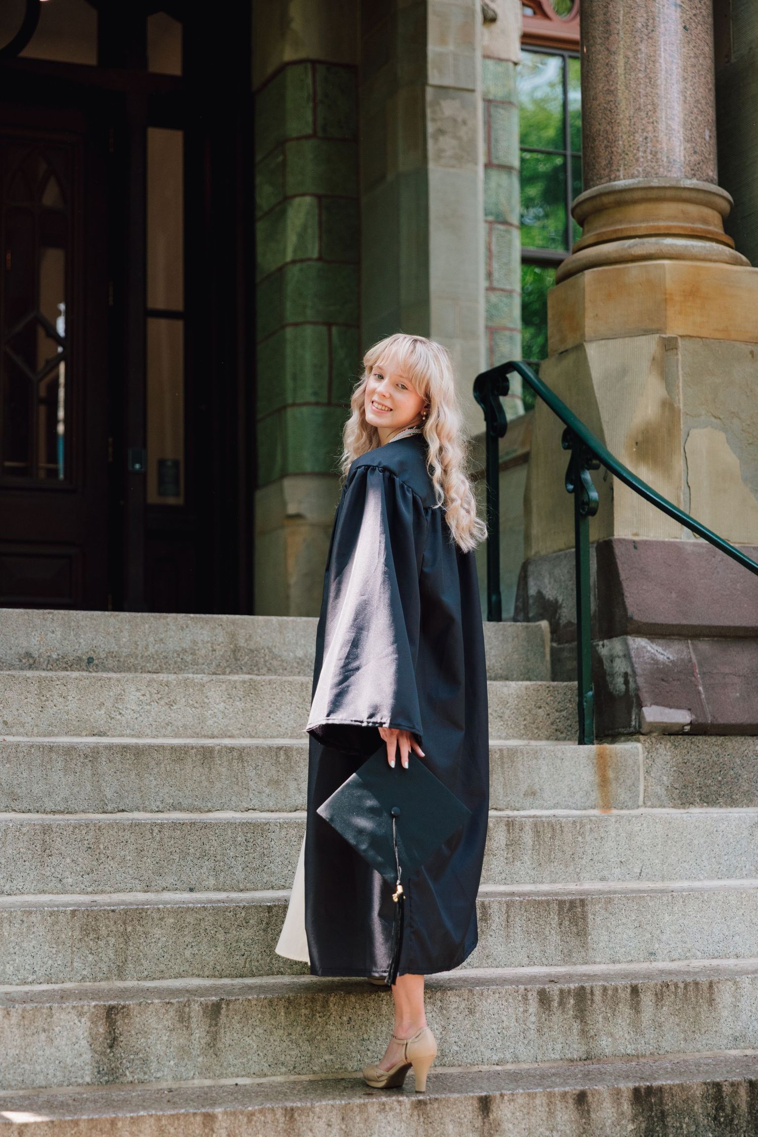 Graduate in academic regalia poses elegantly on stone steps of campus building.