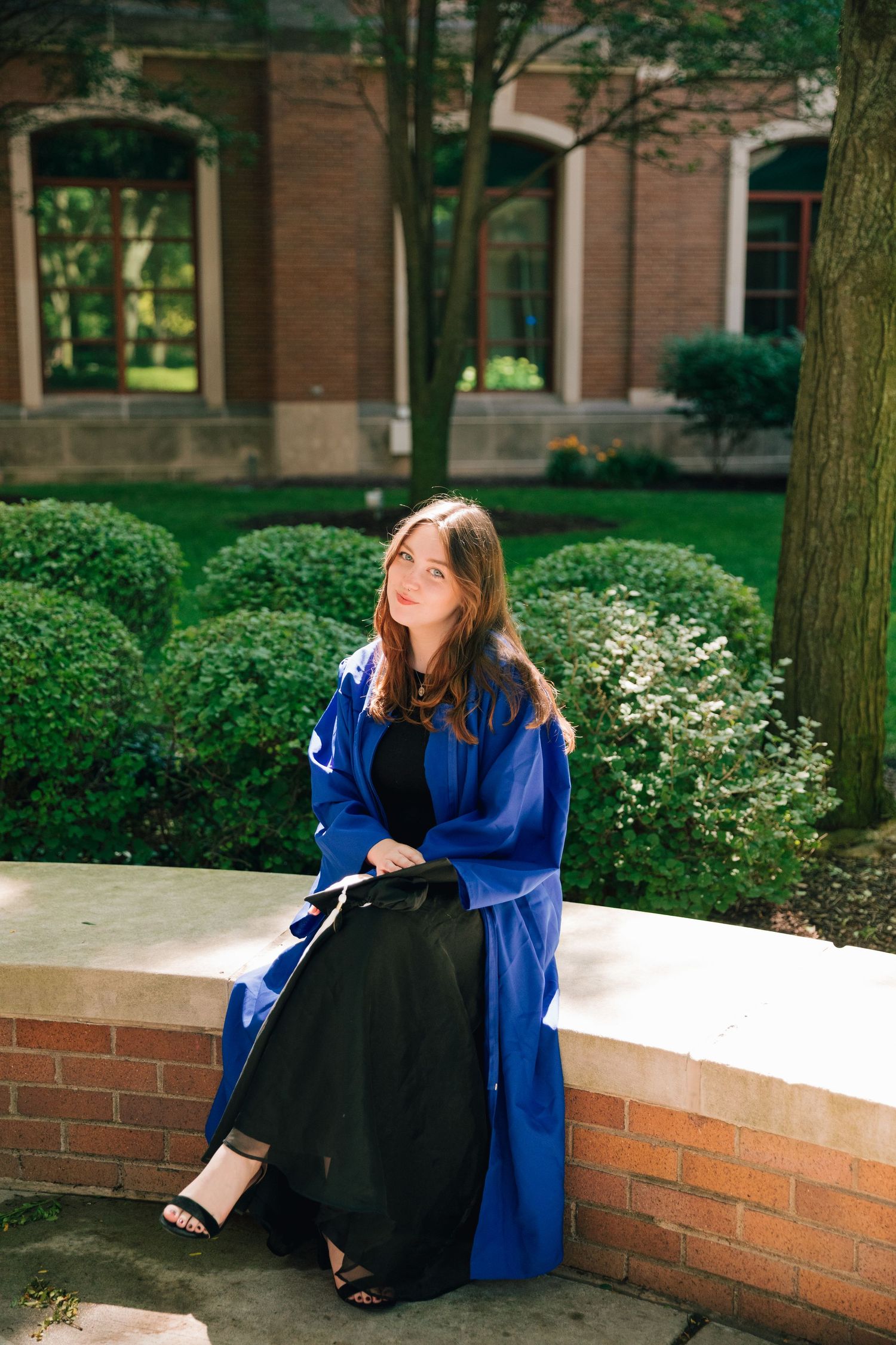 Graduate in blue commencement robe sits on brick wall by landscaped garden on sunny university campus.