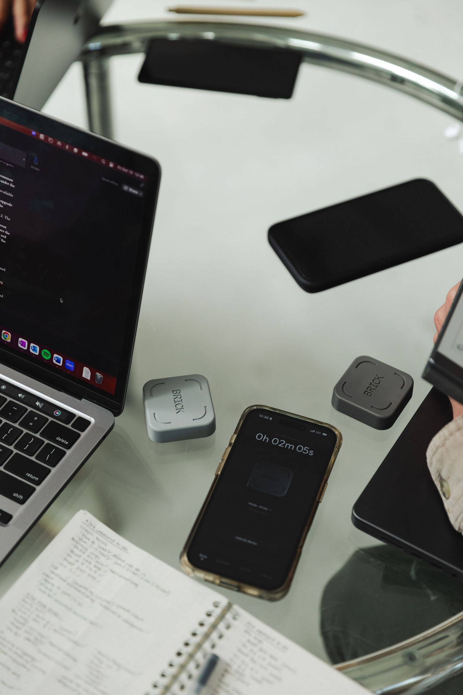Glass desk with MacBook, smartphones, small tech devices, and an open notebook in a modern workspace.