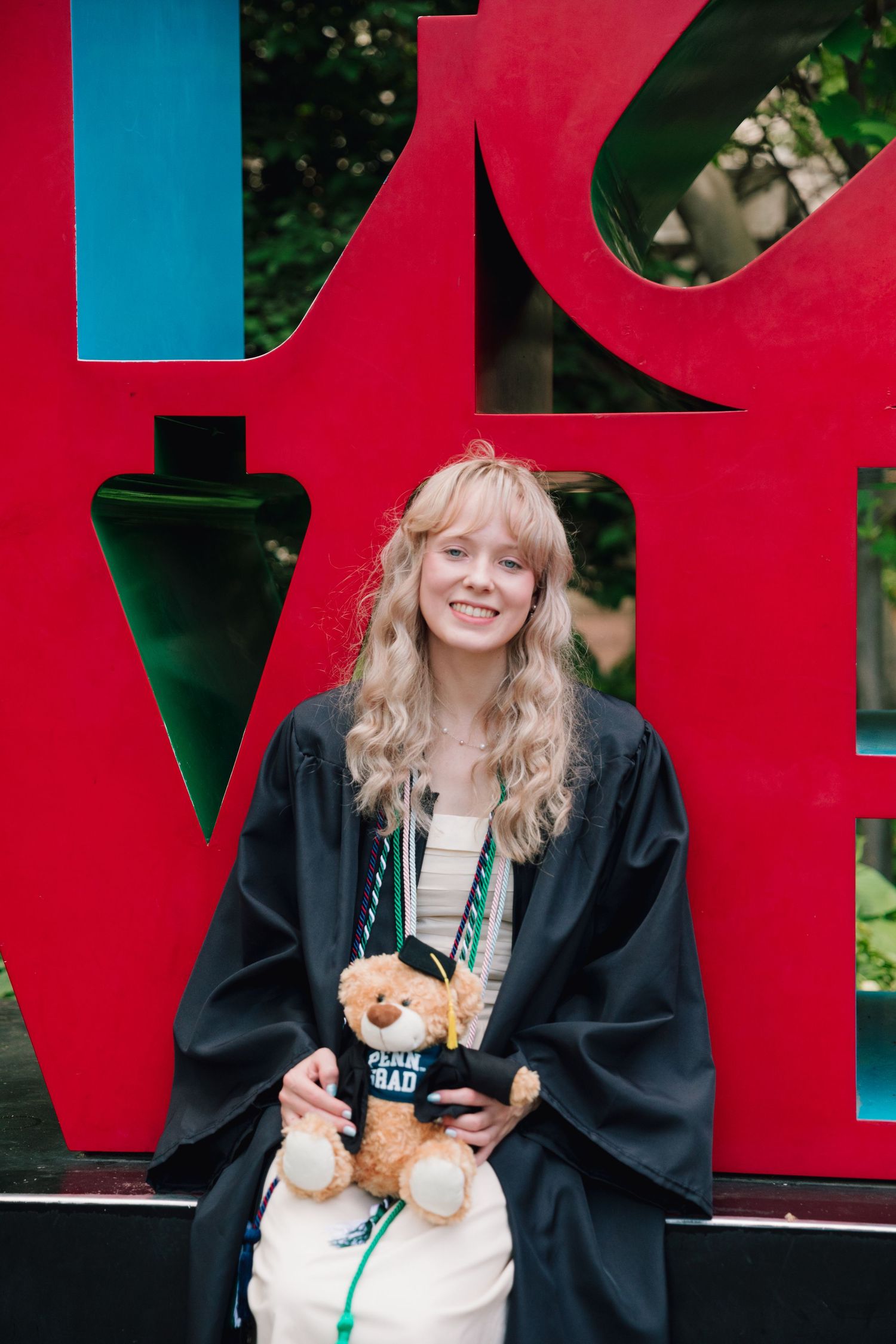 Graduate posing in academic regalia with a teddy bear in front of red LOVE sculpture.
