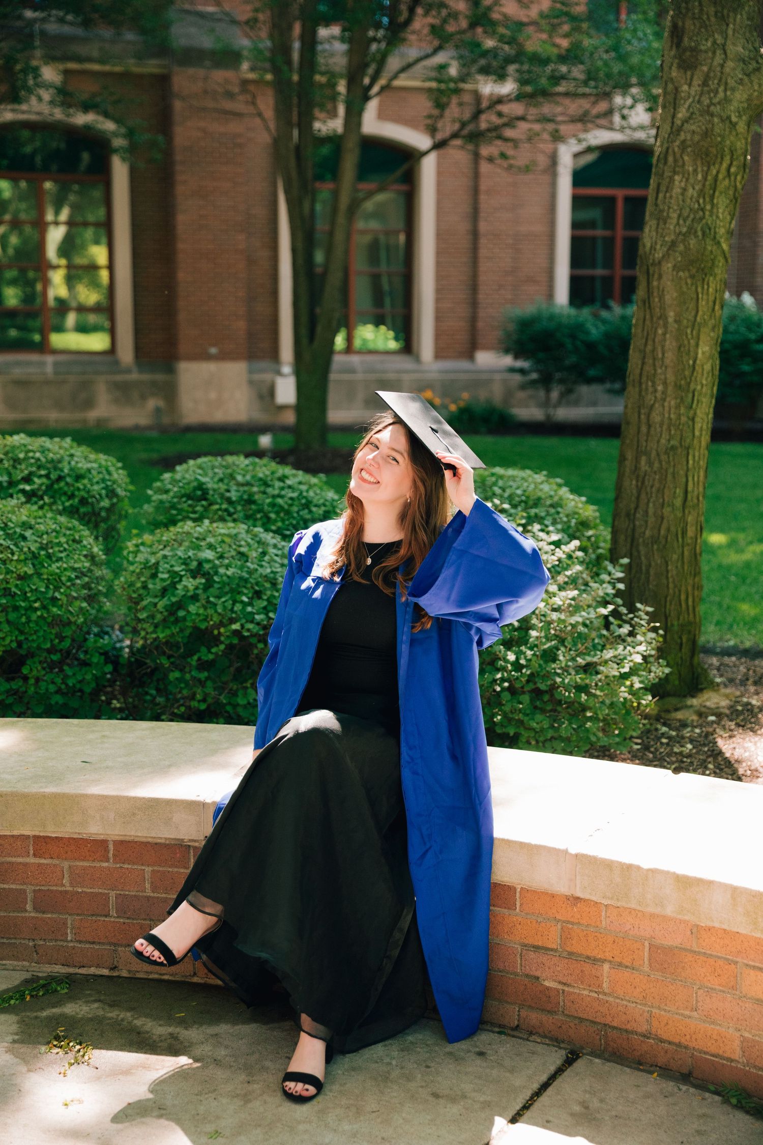 Graduate in royal blue graduation gown relaxes on brick wall during sunny graduation photoshoot.