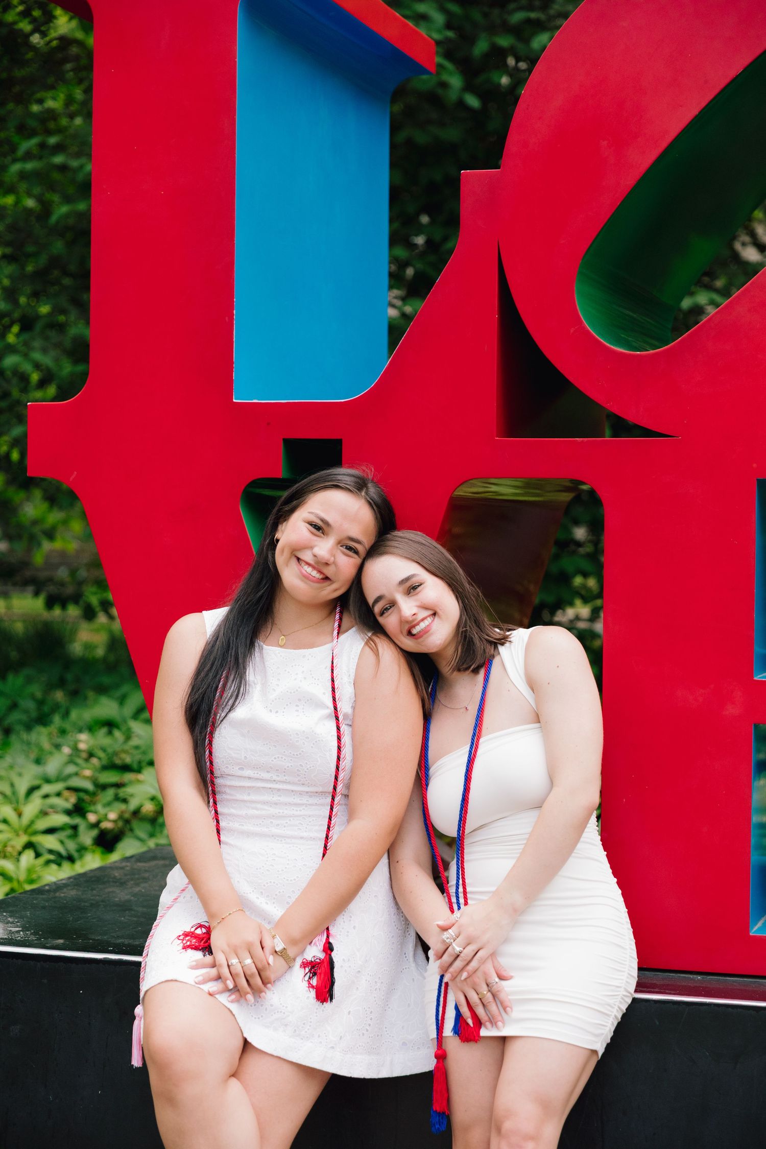 Two graduates in white dresses pose by red LOVE sculpture.