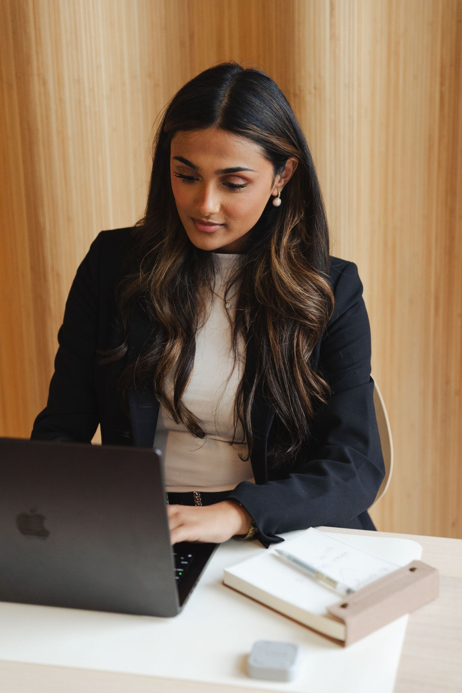Professional woman in black blazer working on Apple laptop at desk with notebook in modern office.