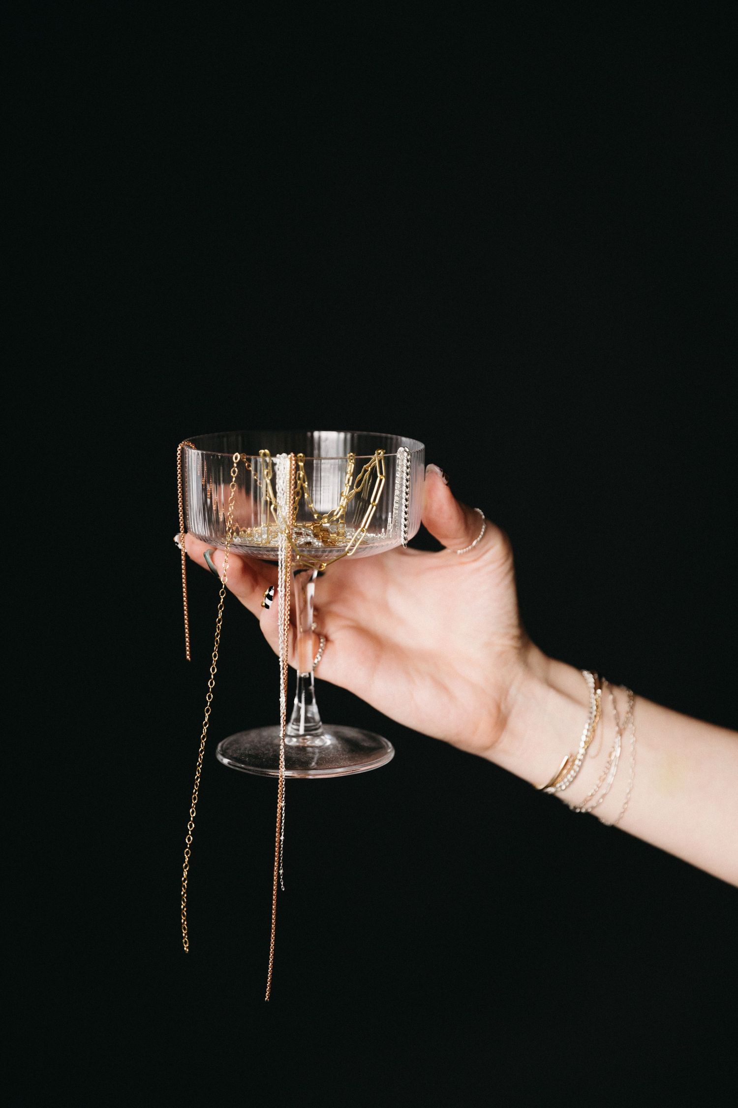 Hand holding a crystal coupe glass filled with gold jewelry and chain earrings against a black background.