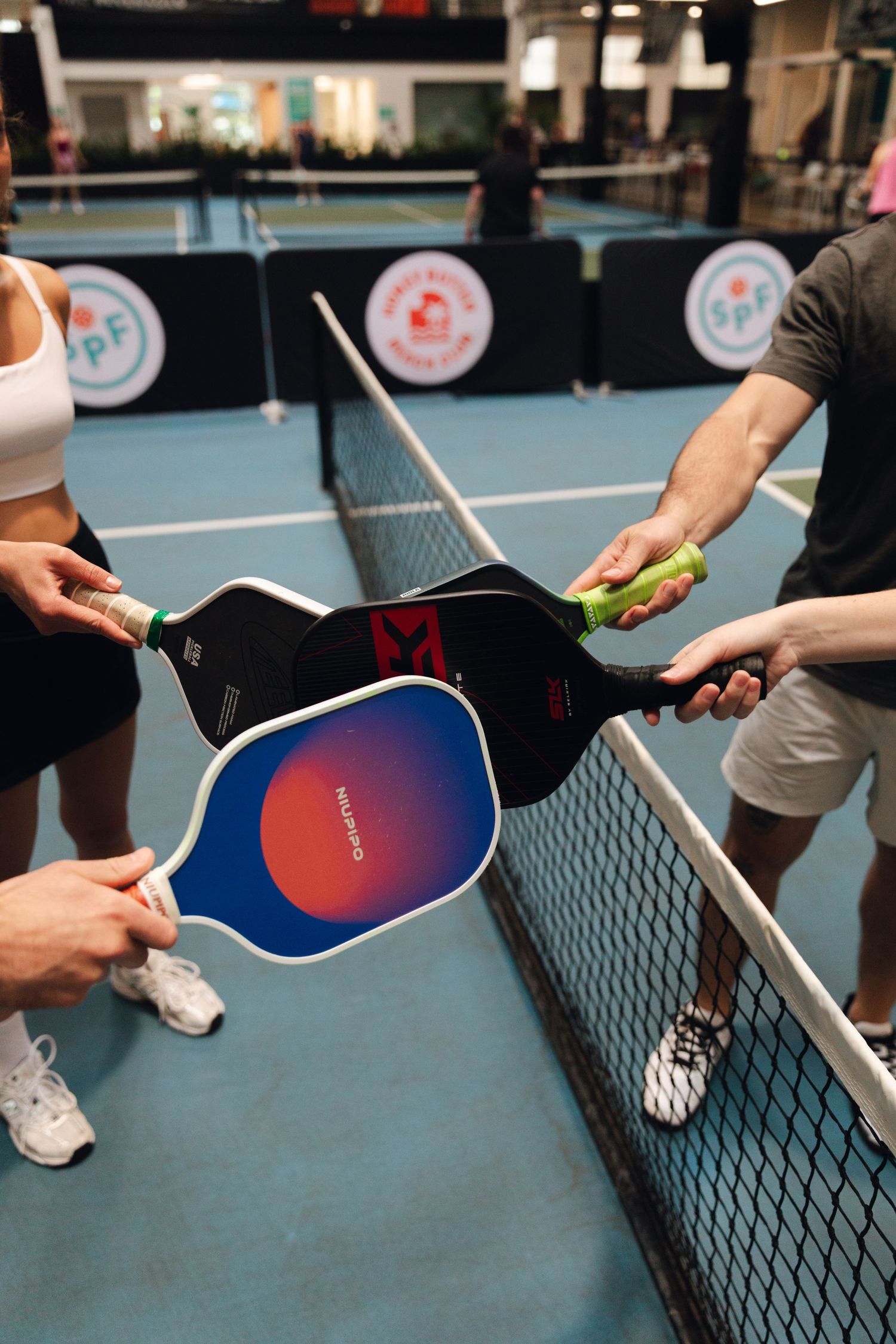 Players tap pickleball paddles over the net at an indoor pickleball court during a friendly post-game handshake.