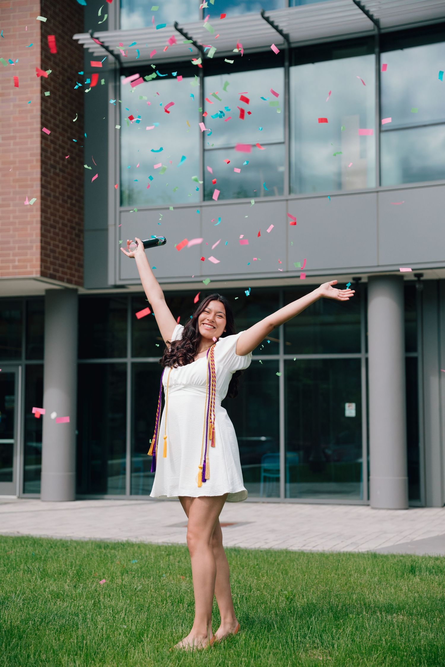 Graduate celebrates with arms raised as pink and blue confetti falls outside university building.