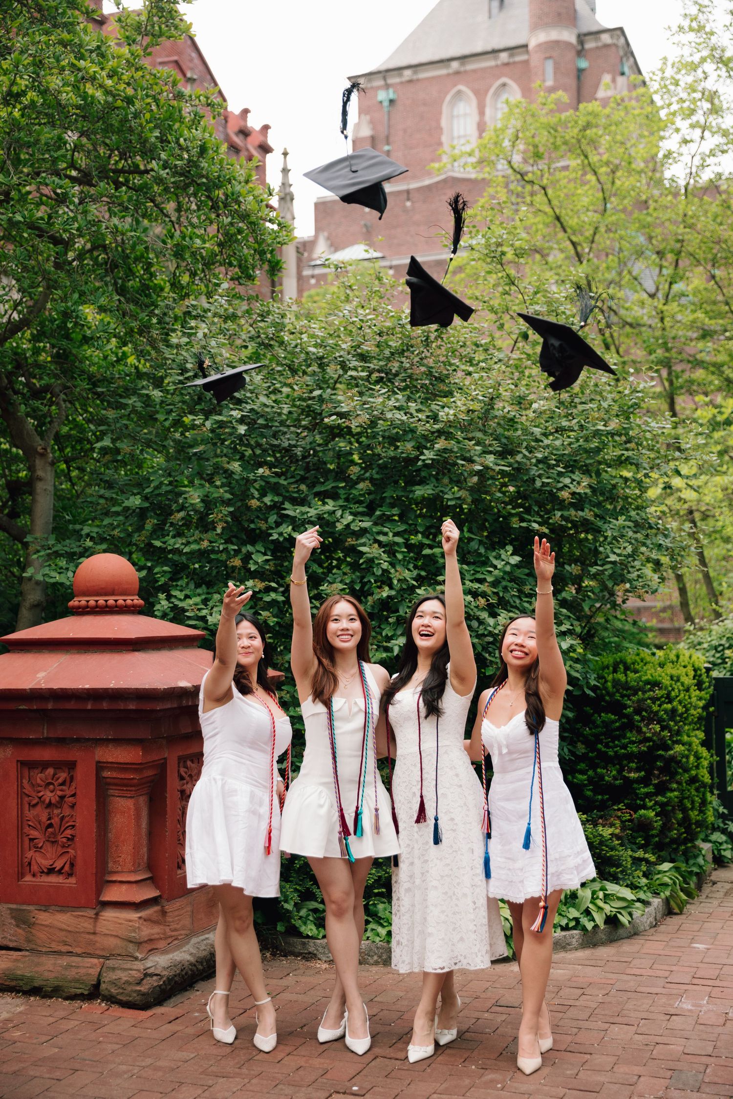 Graduates toss caps in celebration while wearing matching white dresses on campus grounds.