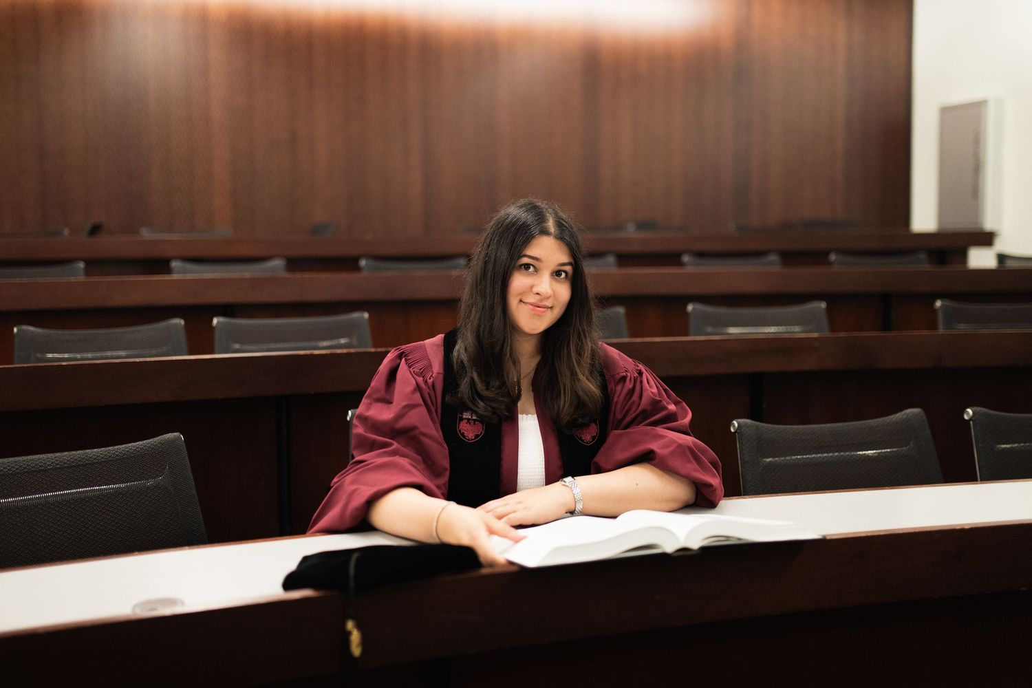 Graduate in maroon regalia studies in lecture hall with wooden desks.
