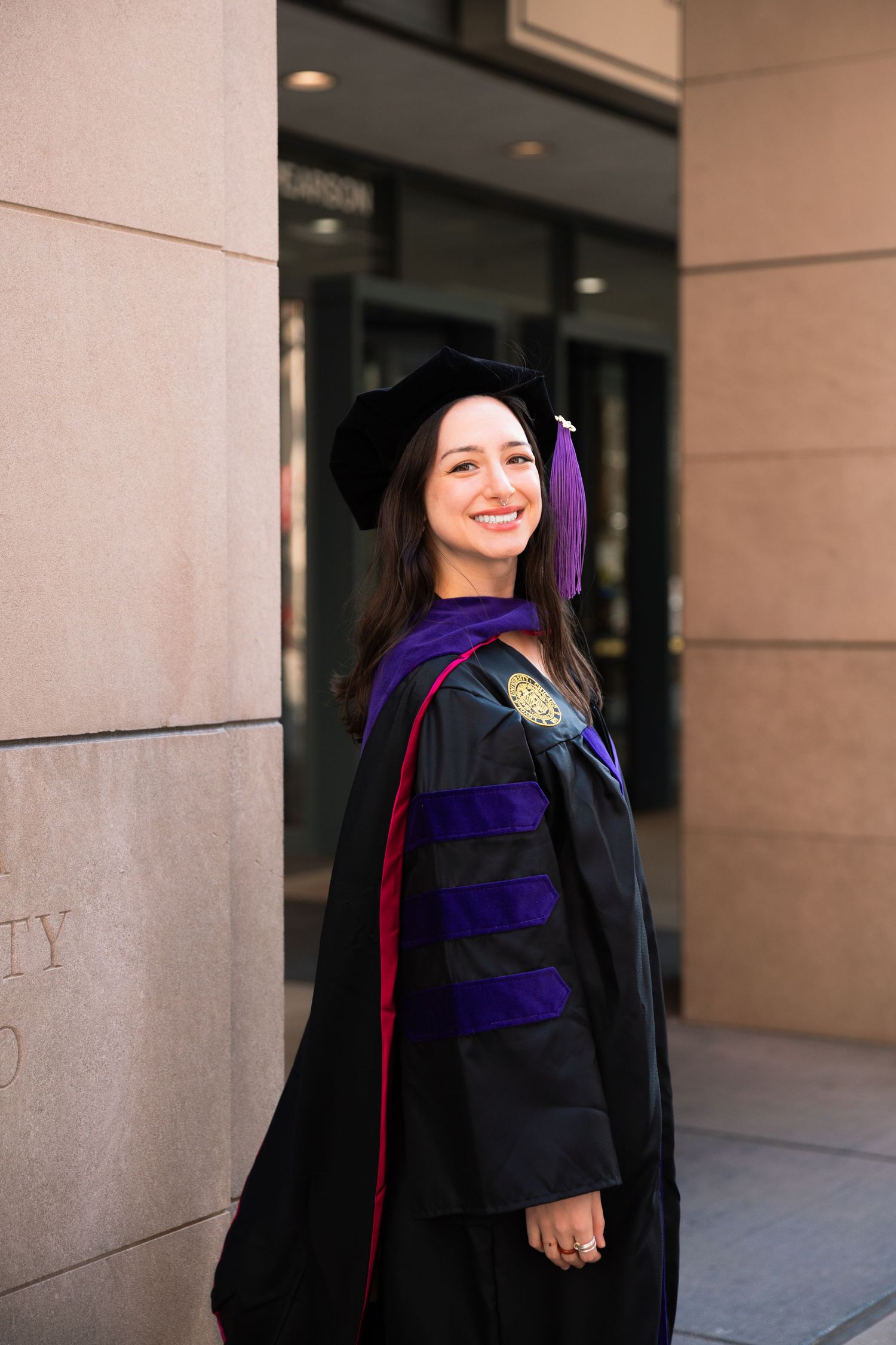 Graduate in academic regalia smiling in front of university building.