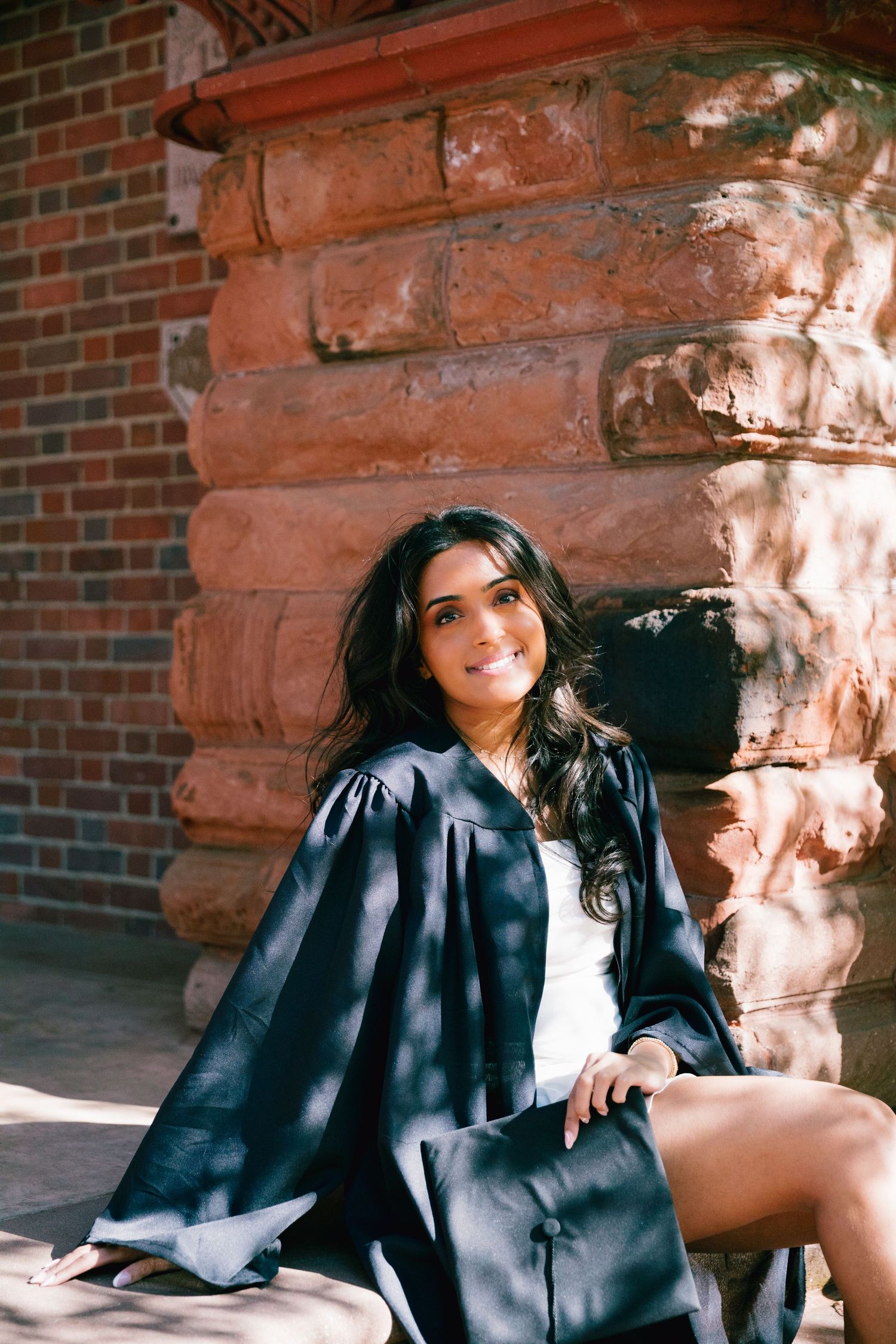 Graduate in academic regalia poses against red brick wall in sunlight.