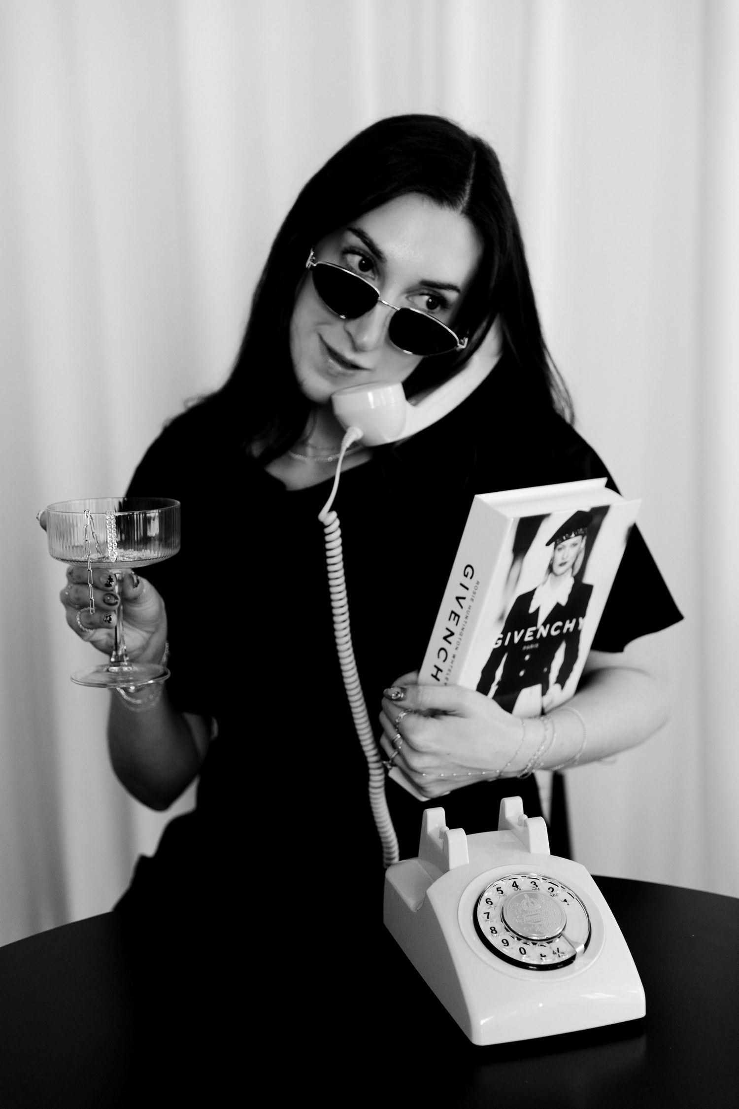 Black and white photo of woman in sunglasses holding retro phone, wine glass, and Givenchy book at table.
