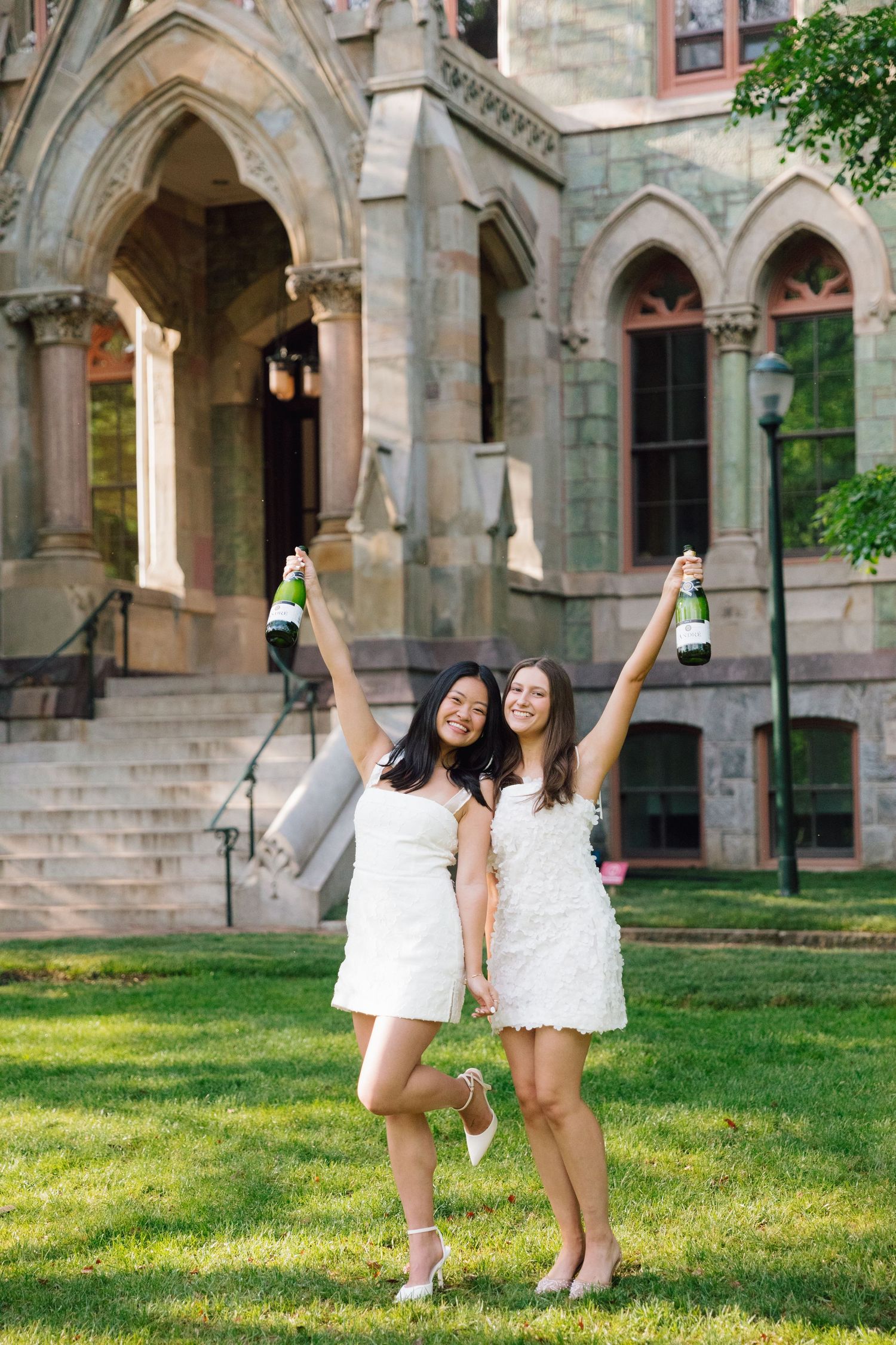 Two graduates celebrate in white dresses on a grassy lawn in front of a historic stone building.
