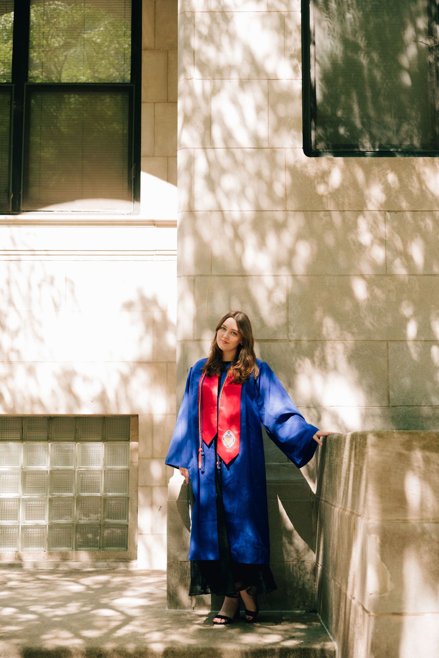 Graduate in bright blue gown poses against stone wall with dappled sunlight.
