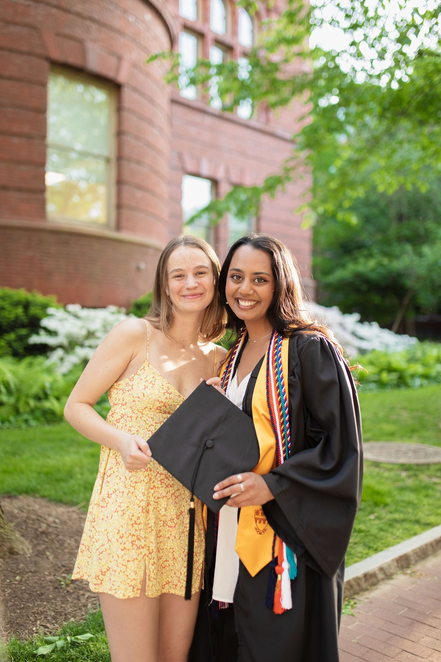 Two graduates celebrate together in front of a brick building on graduation day with cap and gown.