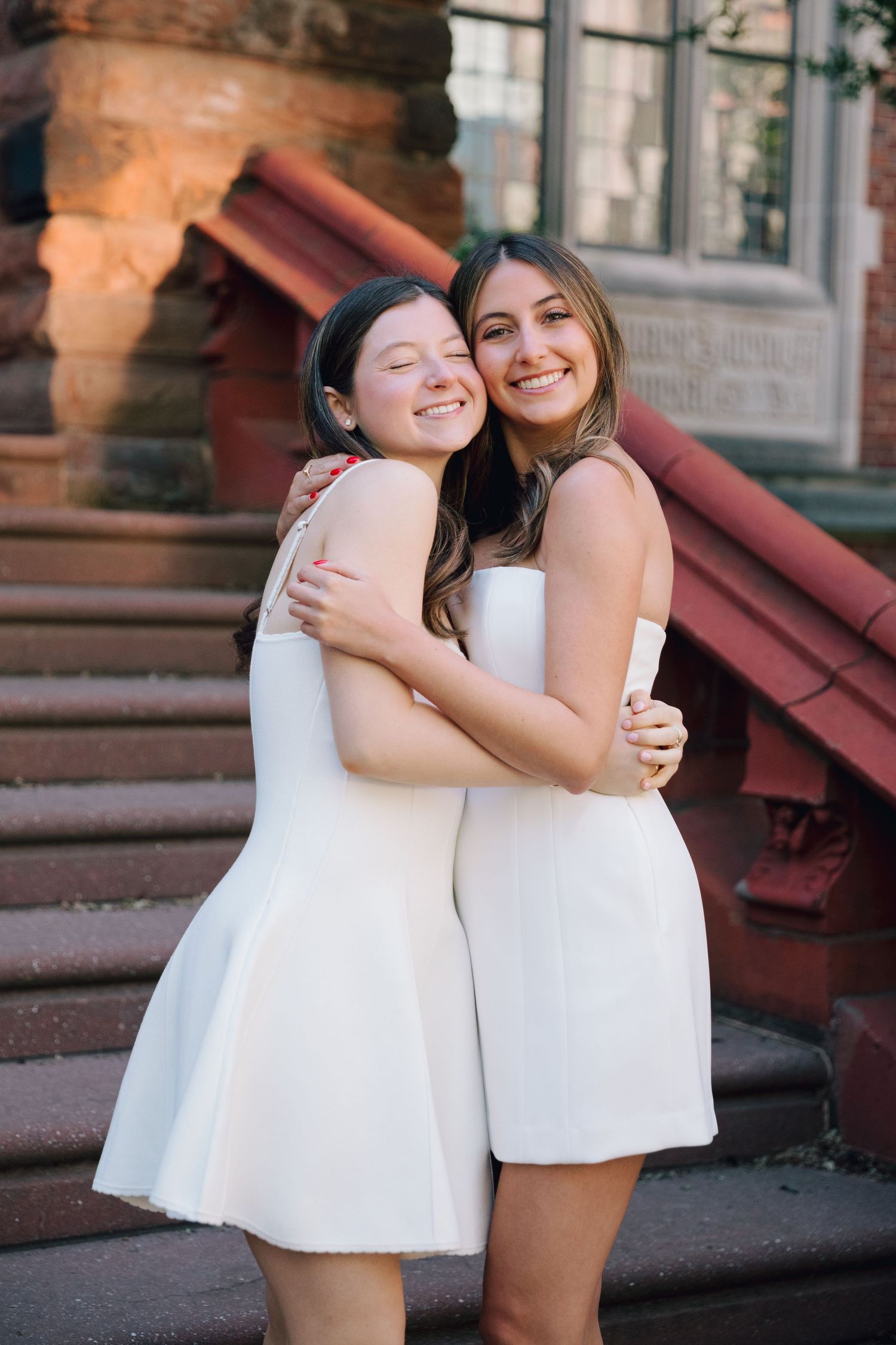 Two people in white dresses embrace and share joyful laughter on brick steps.