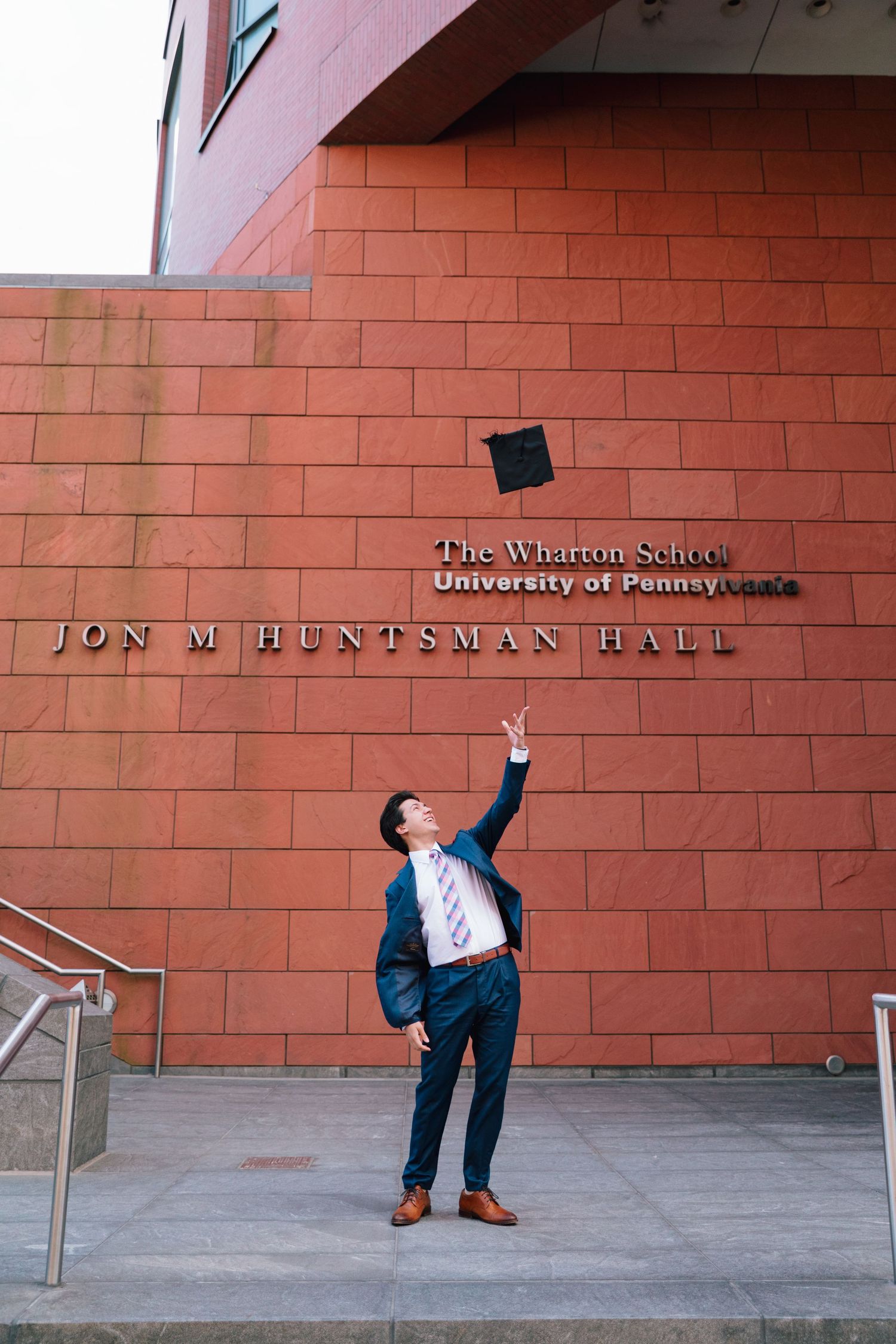 Graduate tosses cap in celebratory poses against red brick wall of Huntsman Hall.
