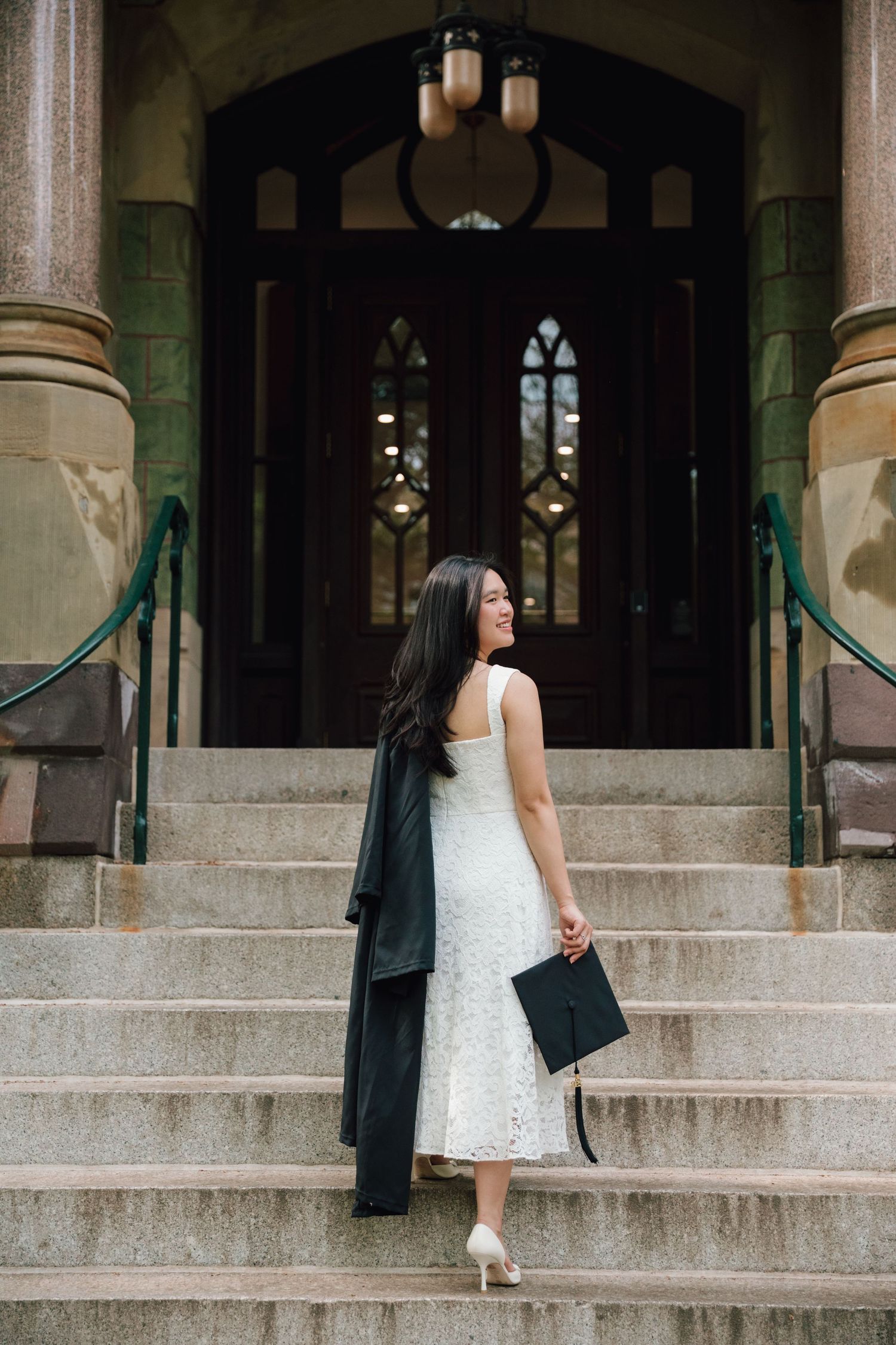 Elegant graduate in white dress poses on historic stone steps in front of Gothic archway.