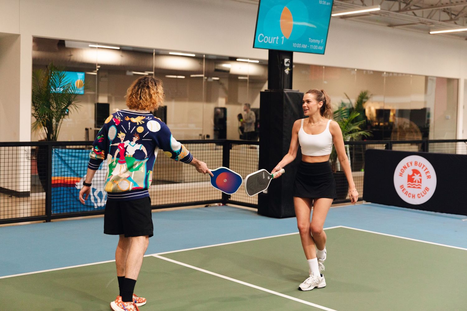 Two women playing pickleball on an indoor court, one in colorful top and one in white crop top and black skirt.