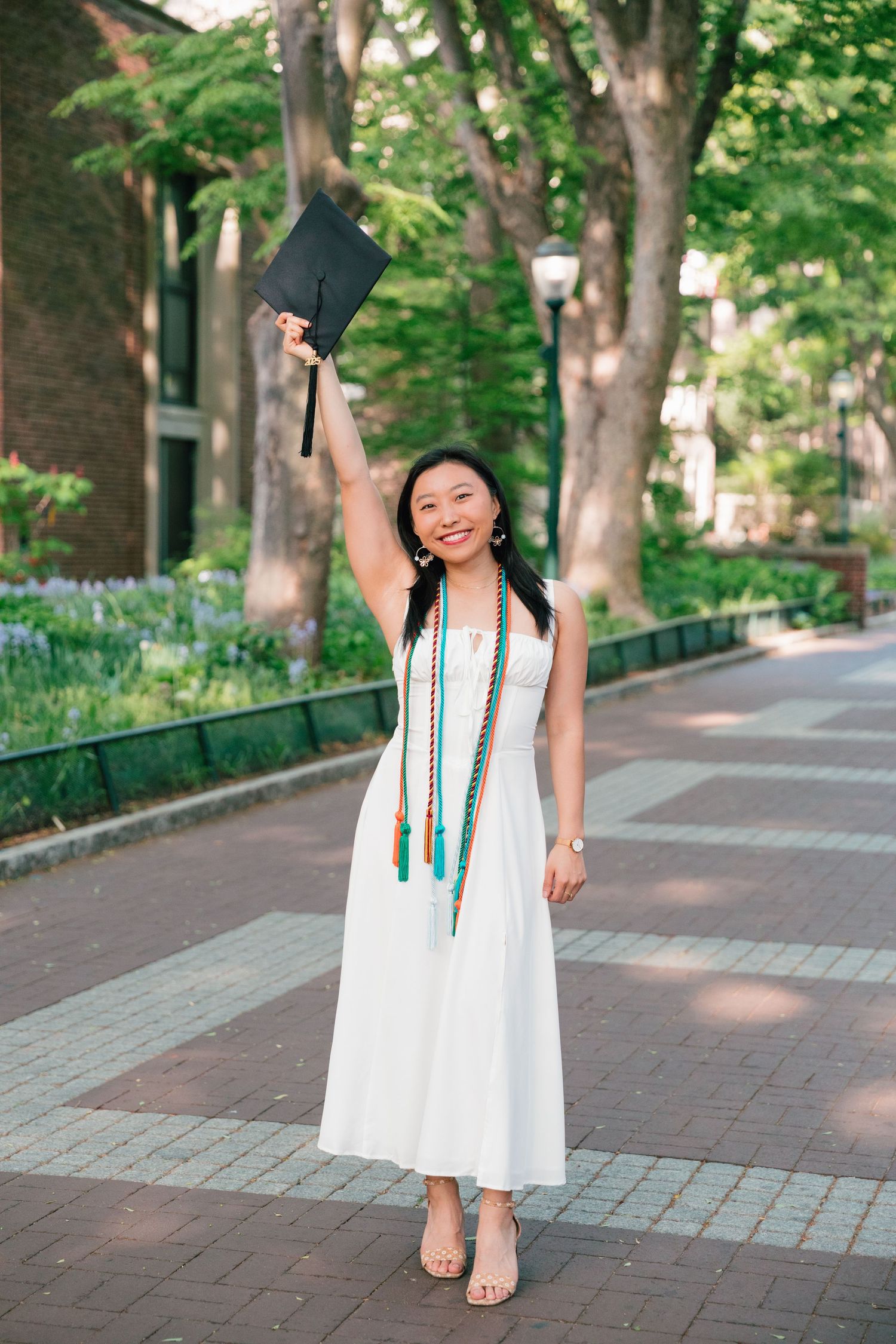 Graduate in white dress with honors cords celebrates with raised cap on campus path.