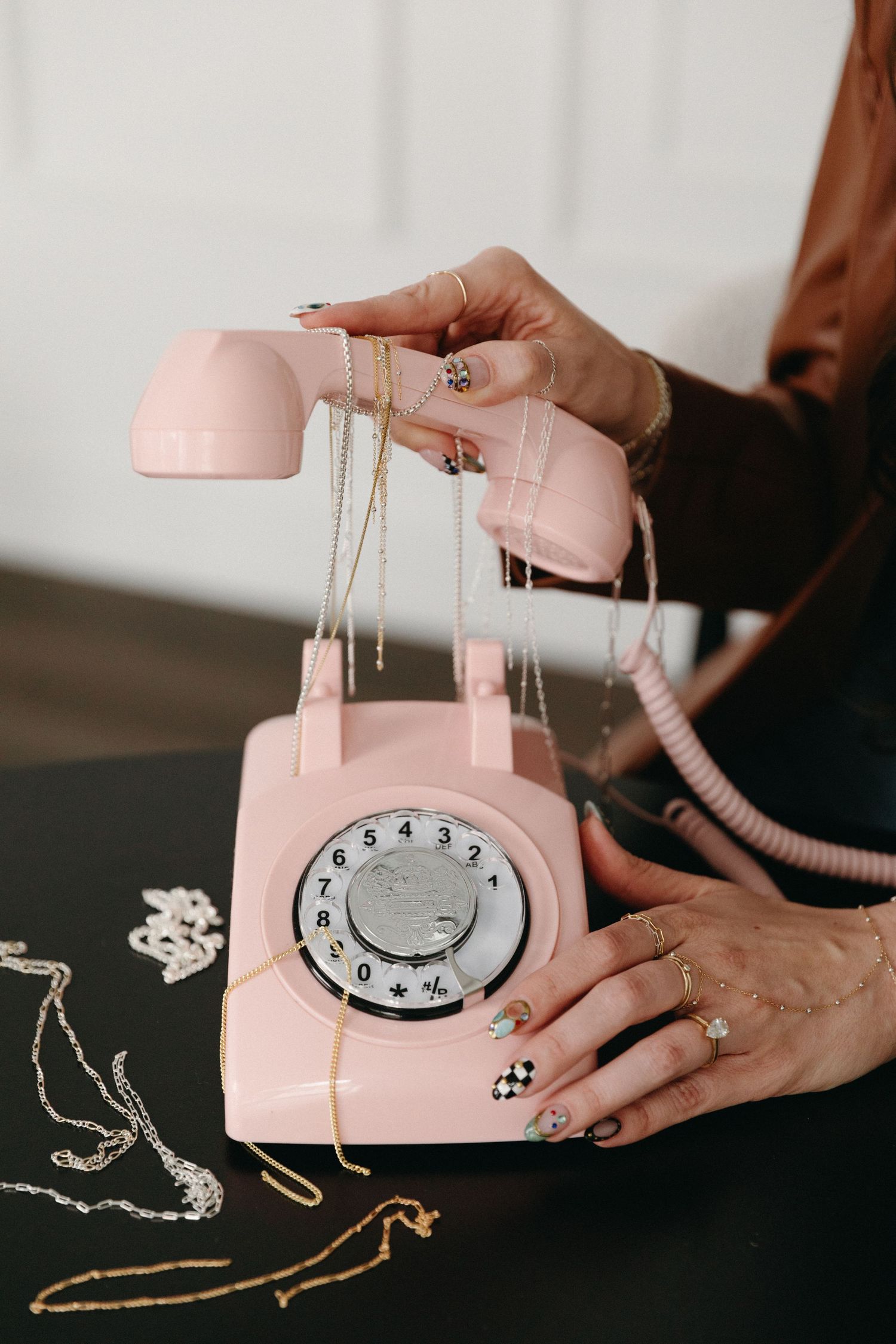 Hands with decorative nails holding gold necklaces over a vintage pink rotary phone on a dark surface.