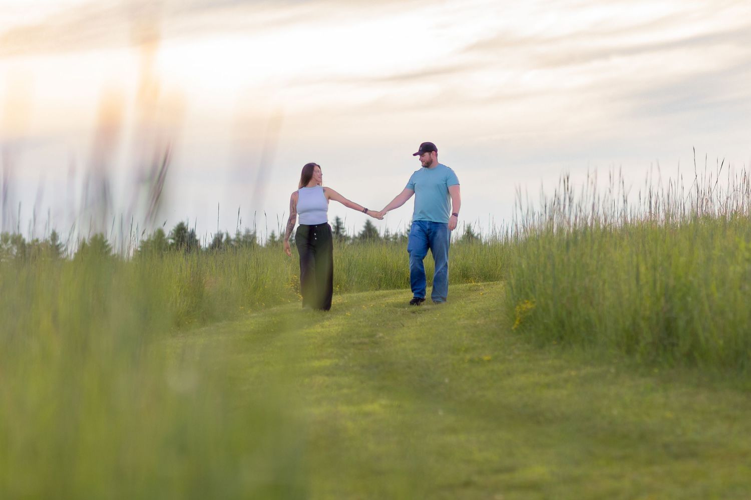 Couple holding hands while walking along grassy path at sunset in rural field.