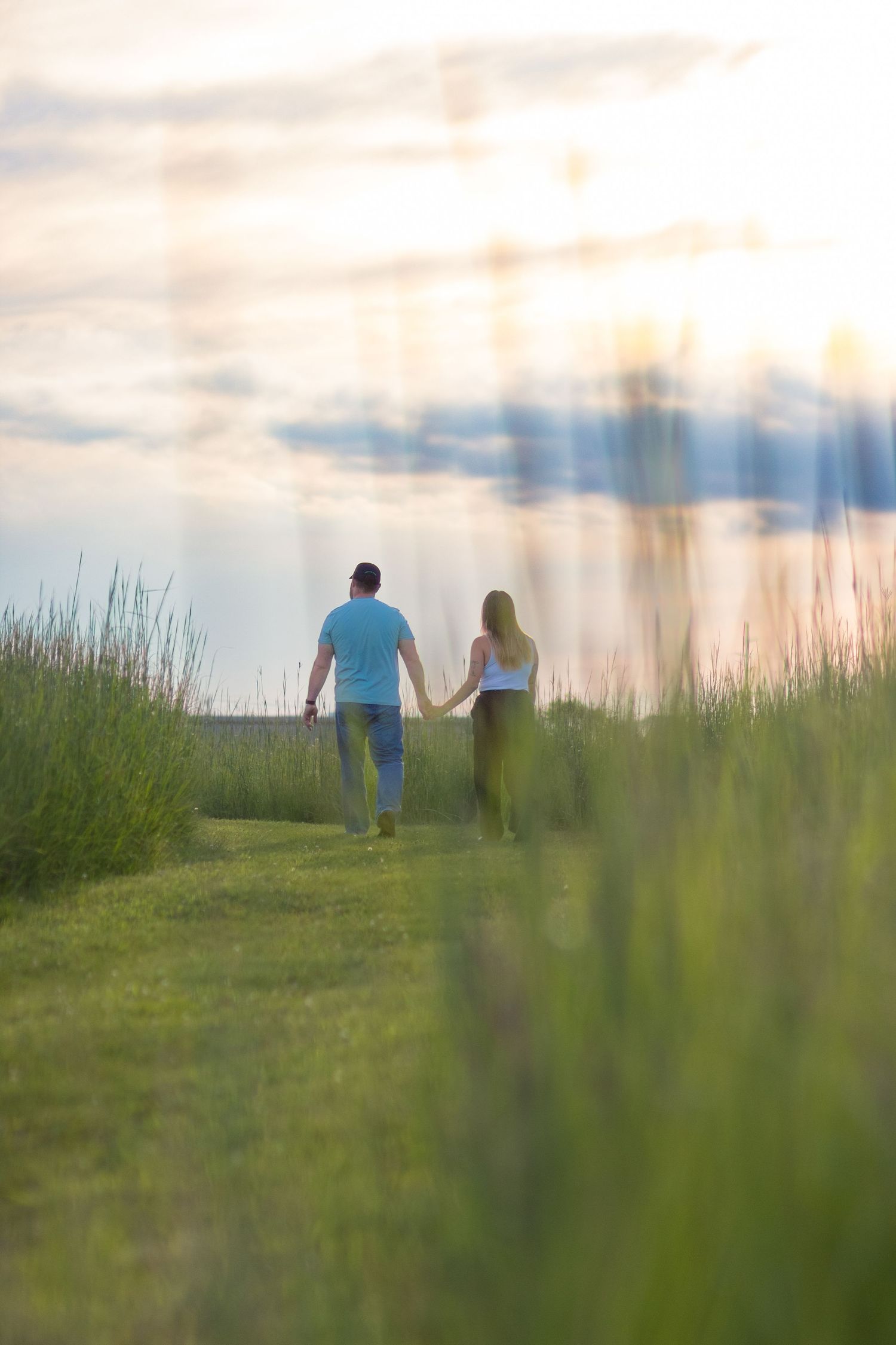 A couple holds hands while walking through a grassy meadow at sunset beside a calm lake.