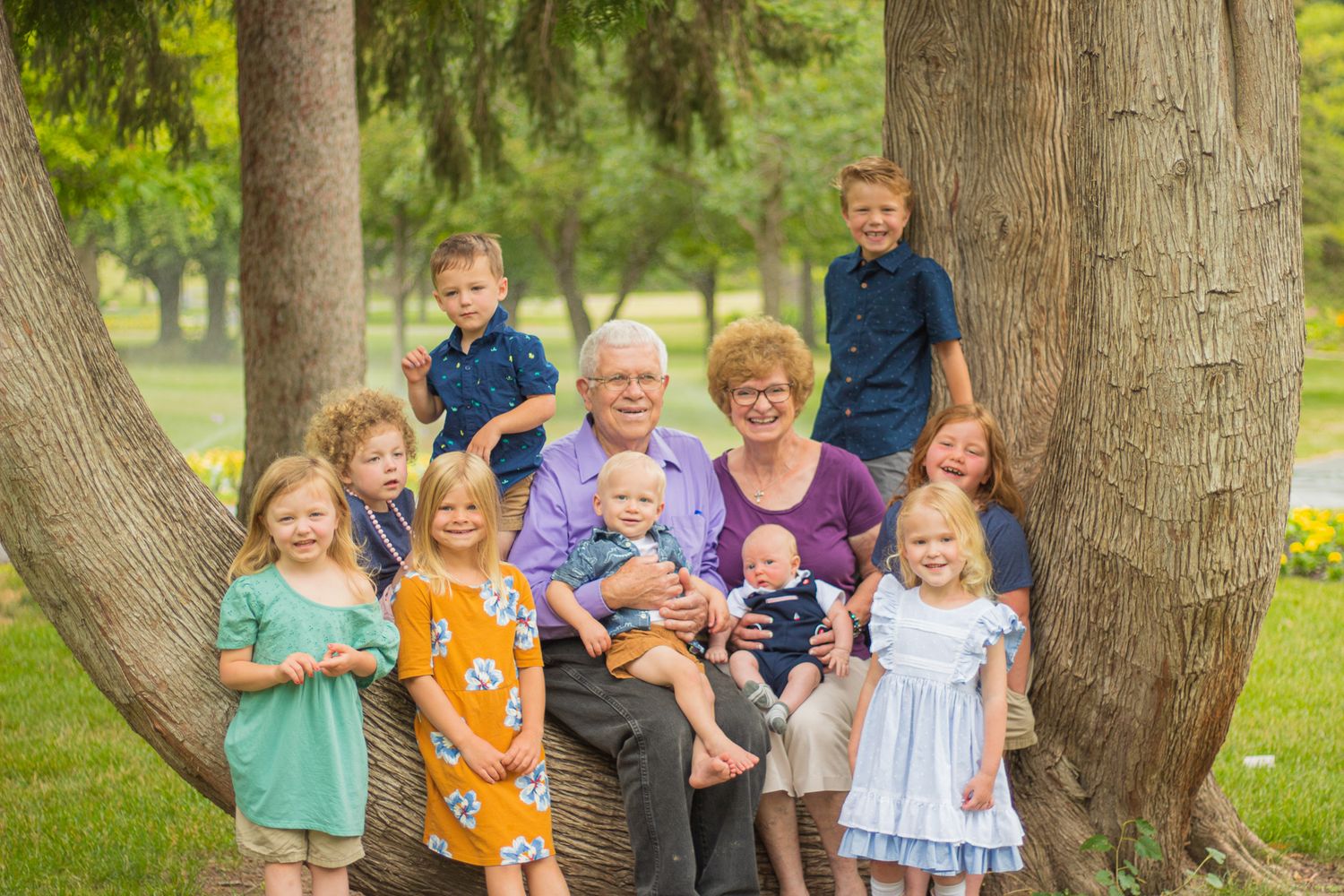 Children and adults gathered around a large tree trunk for a casual outdoor group photo.
